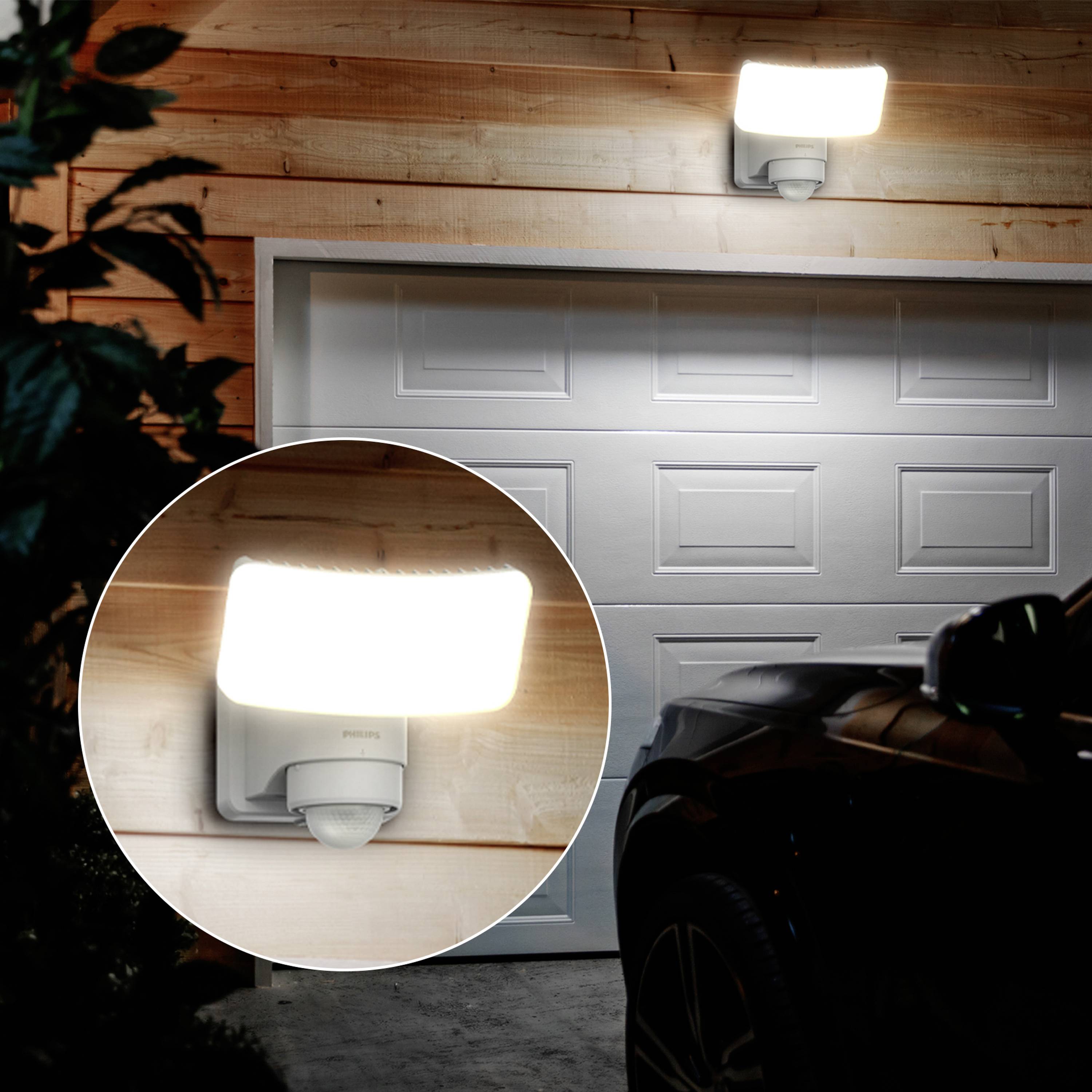 Ein modernes Außenlicht erhellt eine Garage an einem Holzgebäude. Ein Parkplatz mit einem Fahrzeug ist im Vordergrund sichtbar.