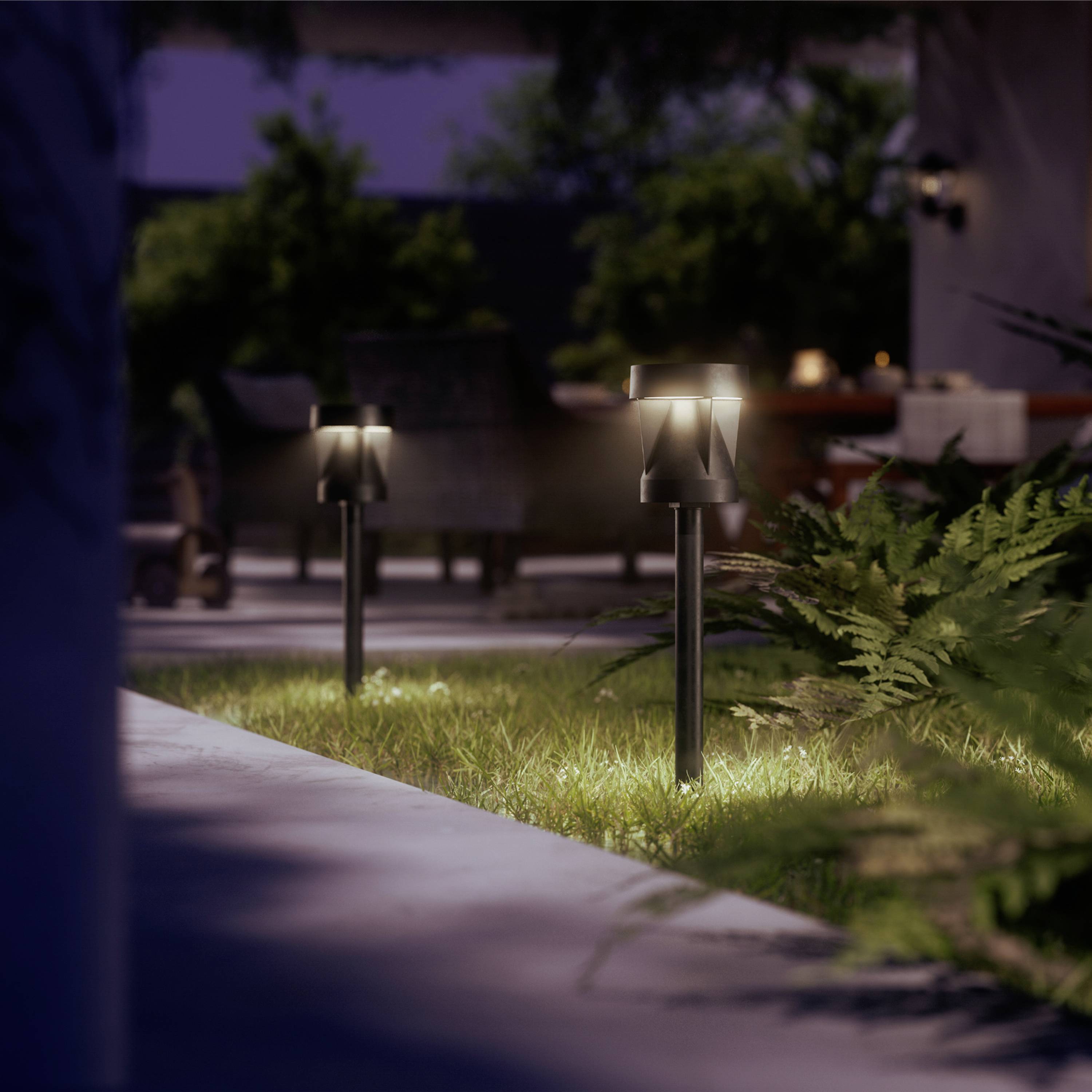 Garten mit beleuchteten Wegleuchten, die den Rasen und Sträucher hervorheben. Im Hintergrund sind Sitzmöbel im Schatten erkennbar.