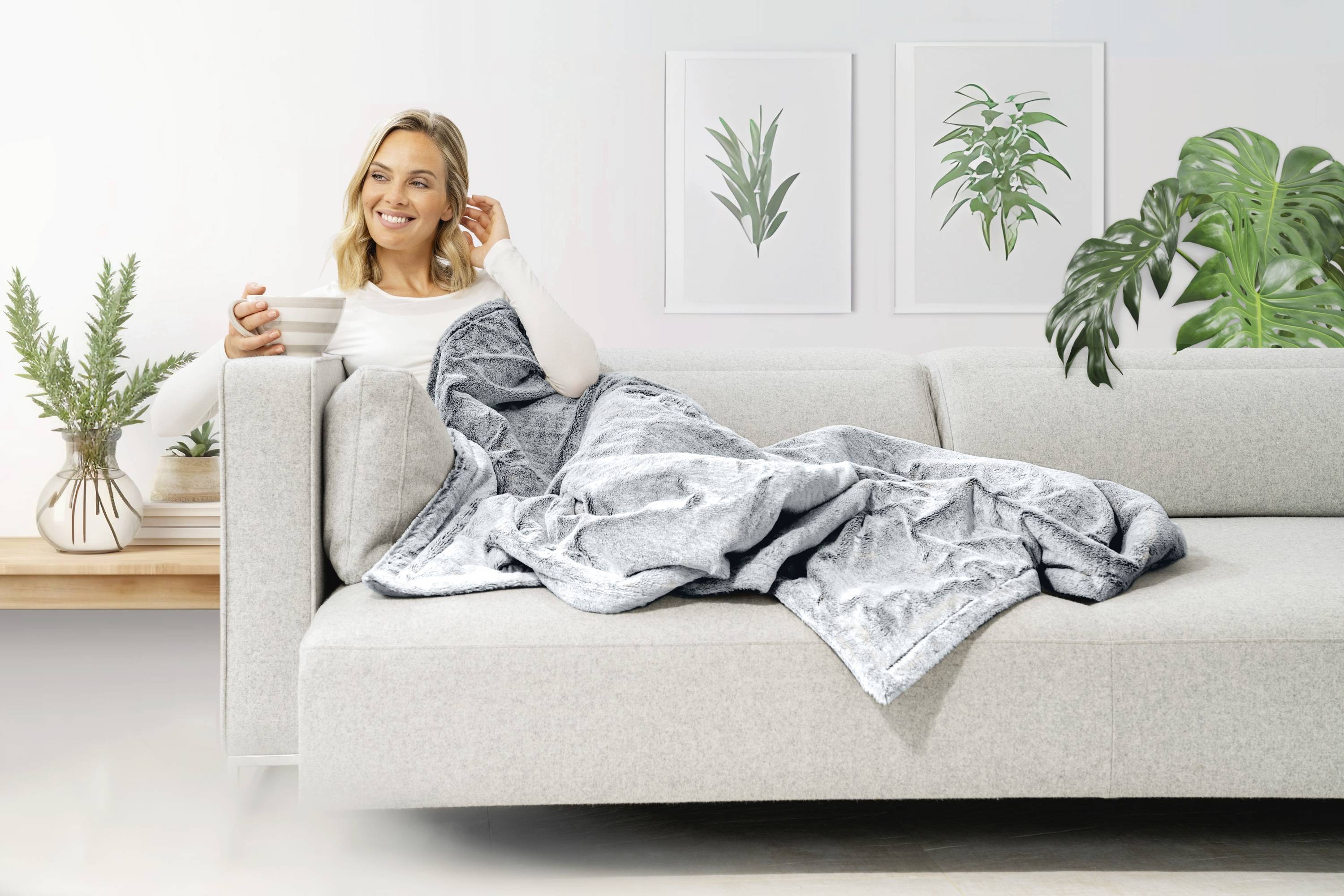 Eine Frau sitzt lächelnd mit einem Getränk auf einem Sofa, umgeben von Pflanzen und in eine graue Decke gehüllt.