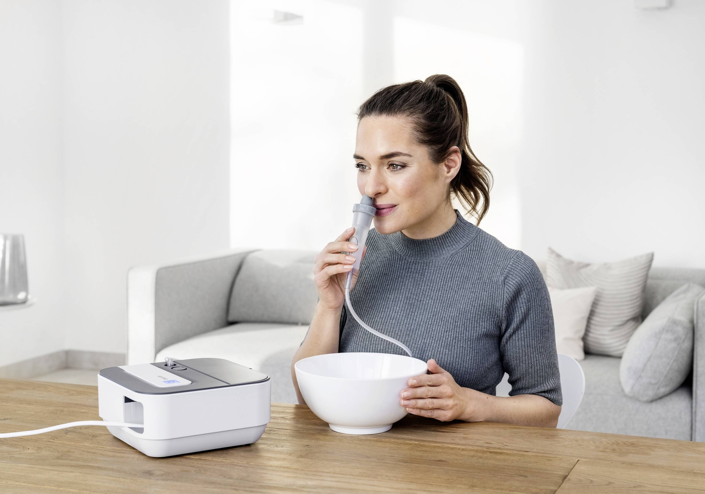 Eine Frau nutzt ein Inhalationsgerät an einem Tisch. Sie hält eine Schale und inhaliert durch einen Schlauch, umgeben von einem hellen Wohnzimmer.