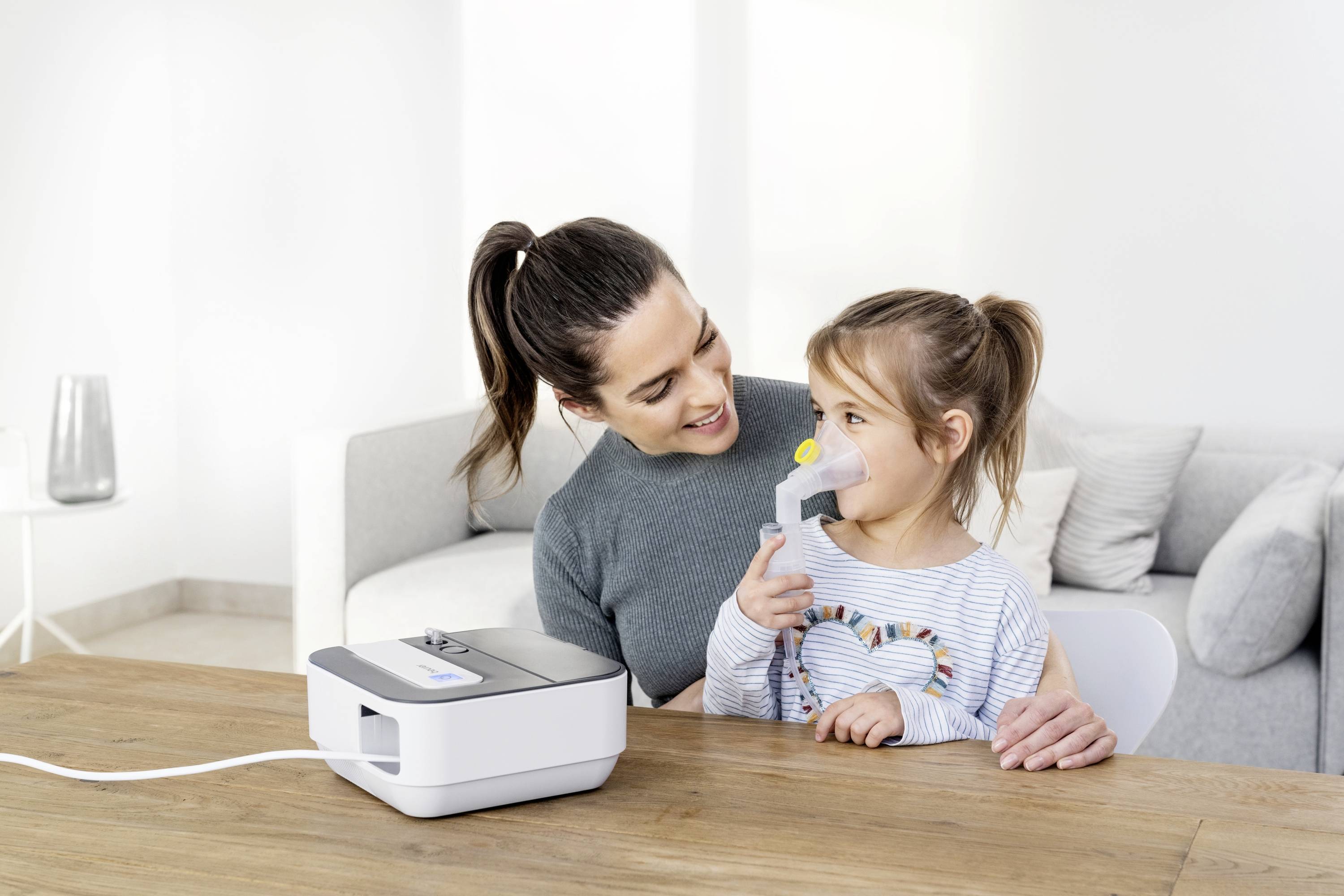 Eine Frau sitzt an einem Tisch und schaut lächelnd ein kleines Mädchen an, das ein Inhalationsgerät benutzt.