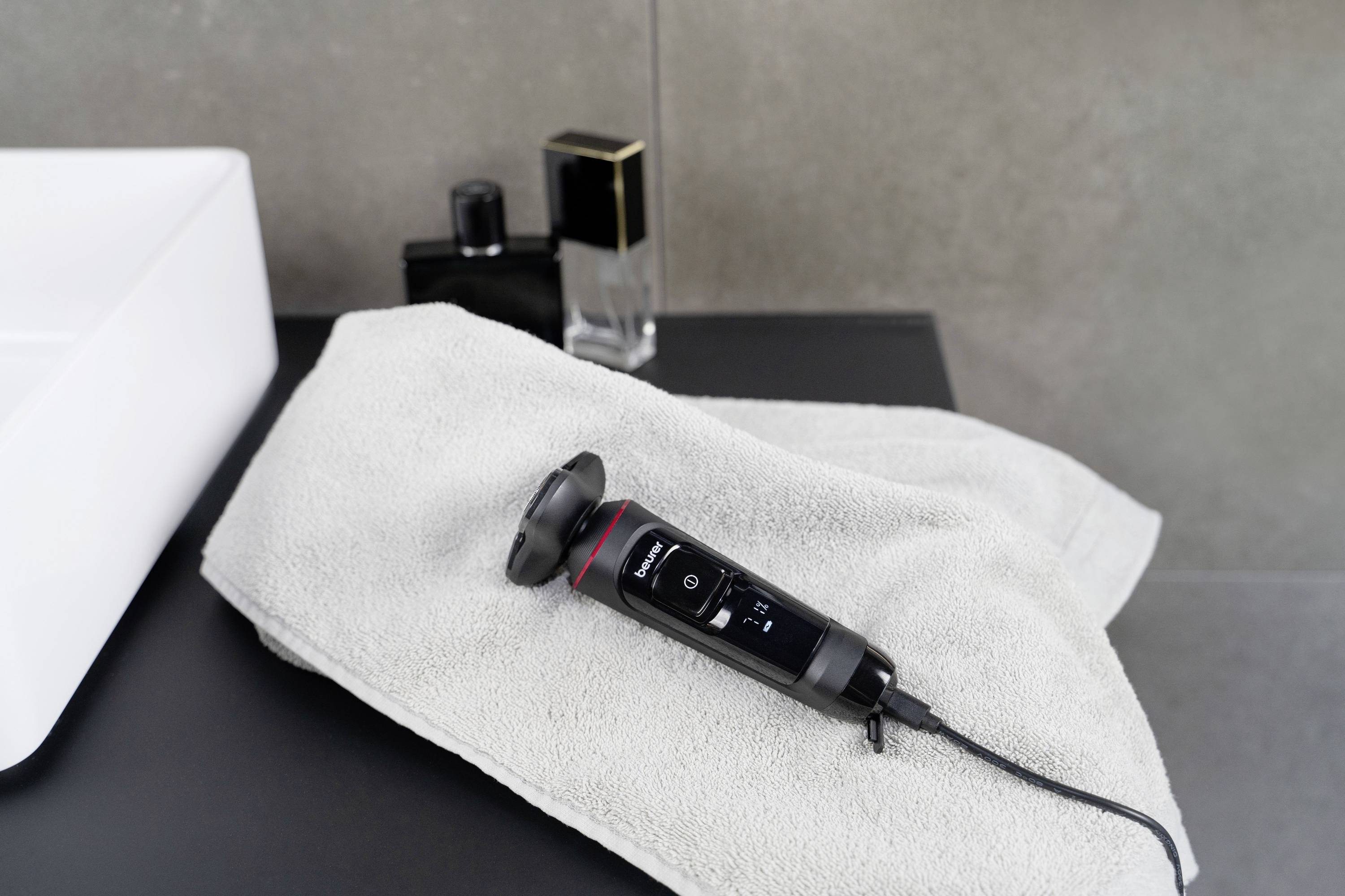 Ein elektrischer Haarschneider liegt auf einem Handtuch auf einem Waschtisch, umgeben von Parfümflaschen im Hintergrund.