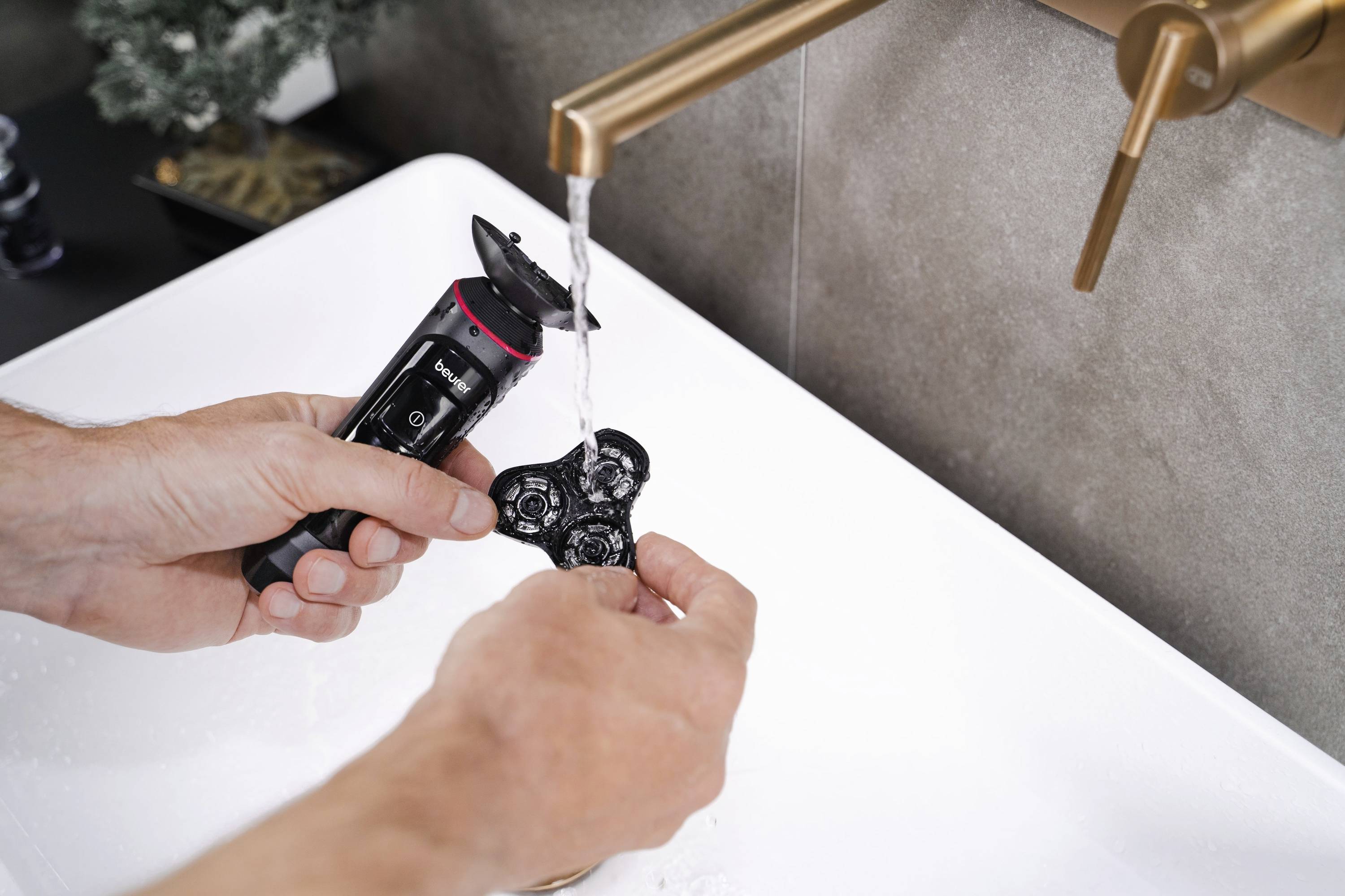 Eine Person wäscht den abnehmbaren Rasierkopf eines elektrischen Rasierers unter fließendem Wasser im Waschbecken.
