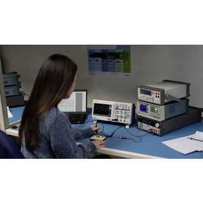 Eine Frau arbeitet an einem Schreibtisch mit einem Laptop und einem Oszilloskop, das an ein elektronisches Gerät angeschlossen ist.