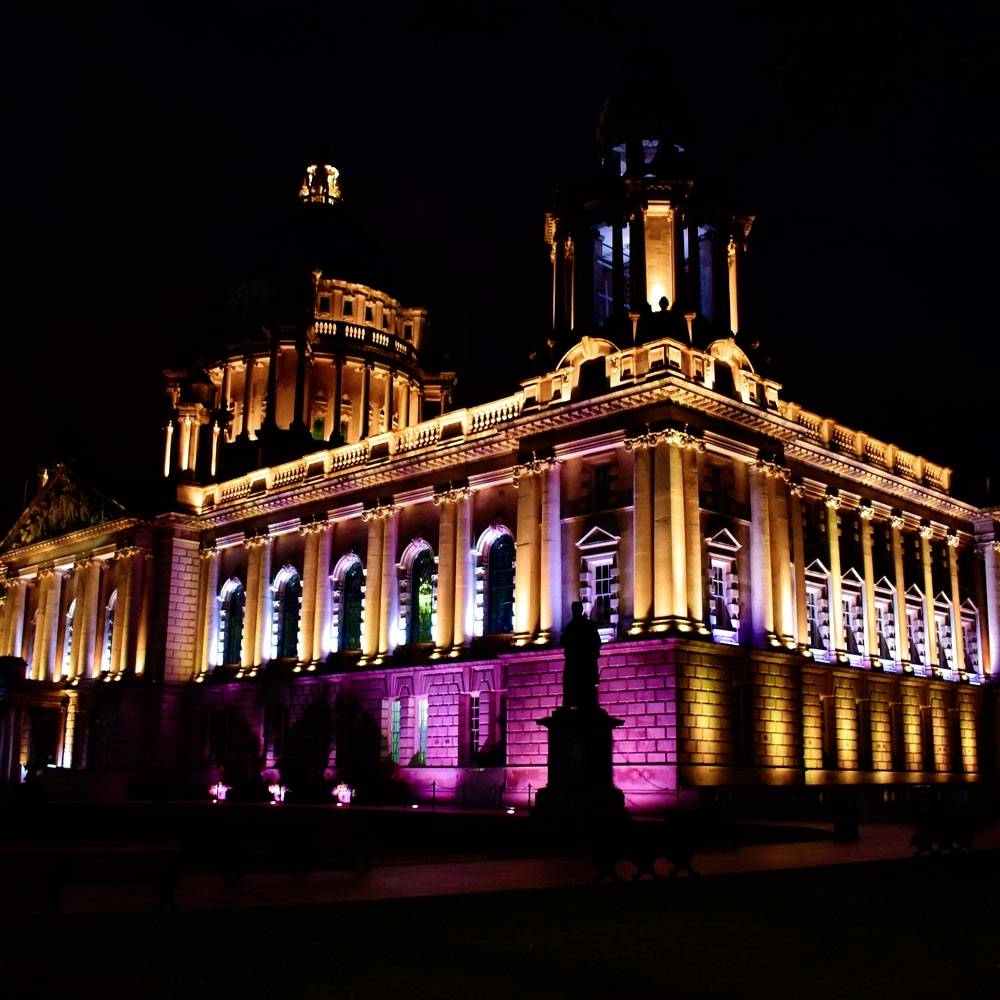 Ein großes, beleuchtetes Gebäude bei Nacht mit pinken und goldenen Lichtern, mit einer Statue im Vordergrund und einem dunklen Himmel.