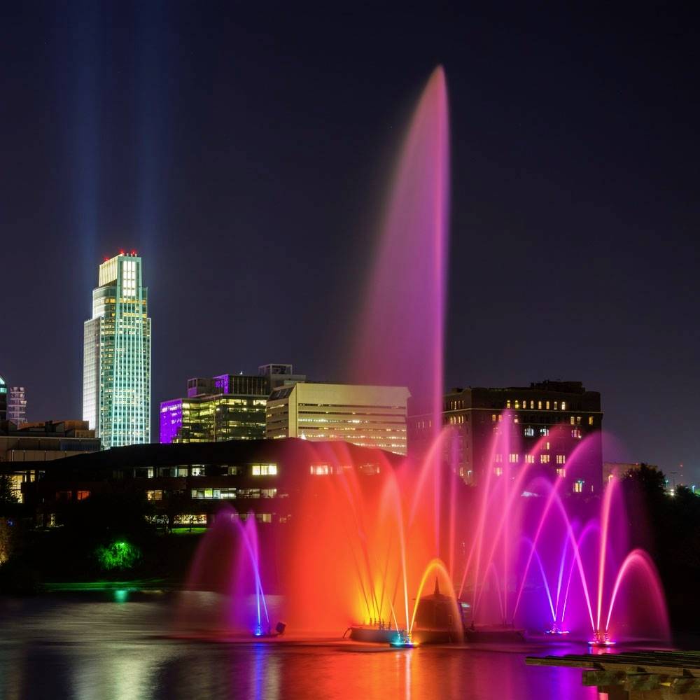 Ein leuchtender Springbrunnen mit bunten Lichtern in einer nächtlichen Stadtlandschaft, Hochhäuser im Hintergrund.