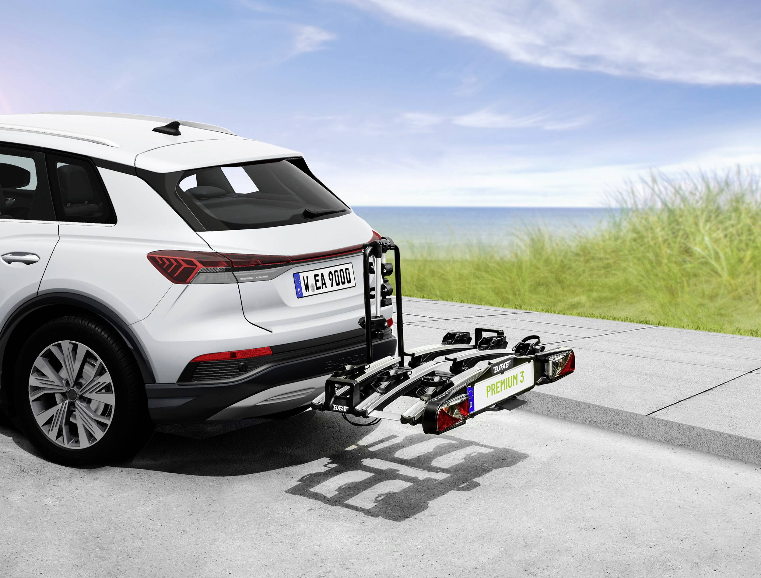 Ein weißes Auto mit einem montierten Fahrradträger am Heck steht auf einer Straße mit einer Graslandschaft und blauem Himmel im Hintergrund.
