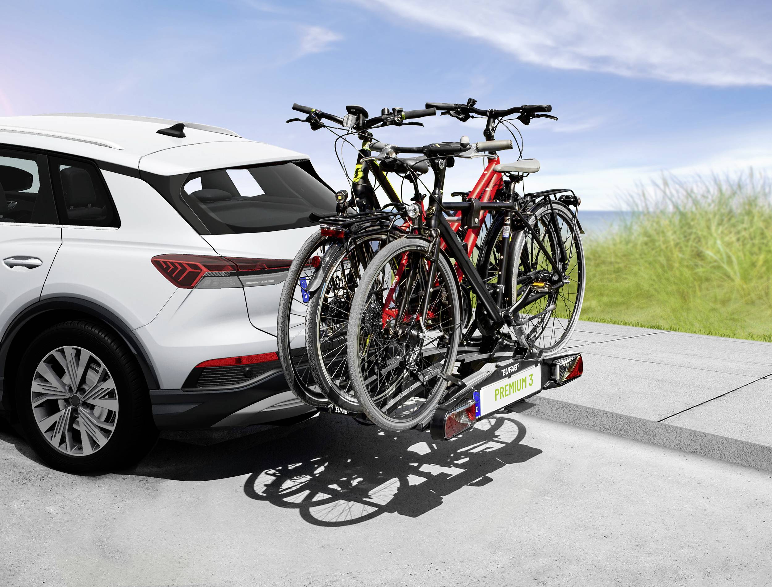 Ein weißes Auto mit einem Fahrradträger, der vier Fahrräder transportiert, steht auf einer Straße. Im Hintergrund sind Gras und blauer Himmel zu sehen.