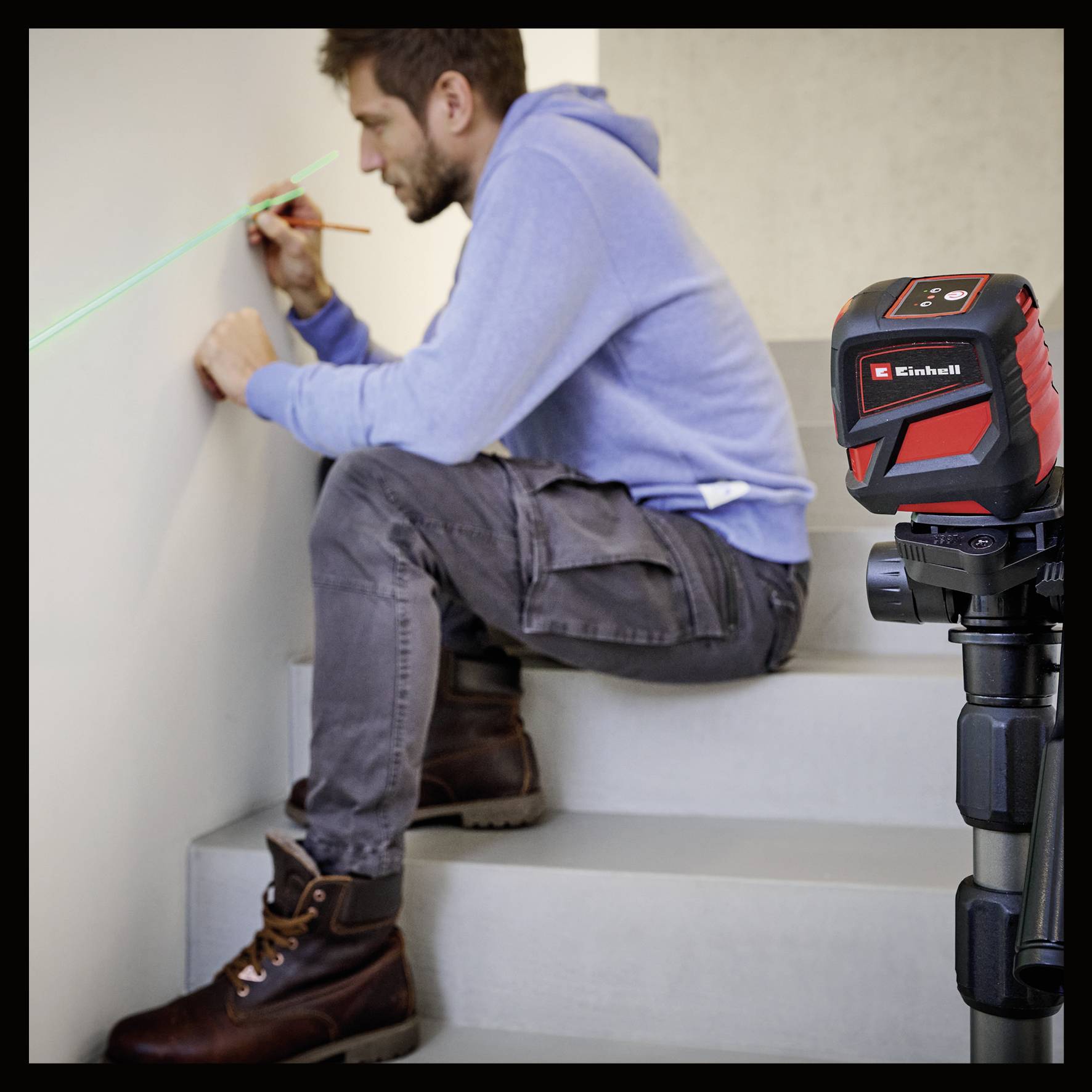Mann in Arbeitskleidung nutzt Laser-Wasserwaage auf Stativ, zeichnet Linie an Wand. Werkzeuge für präzise Messarbeiten.