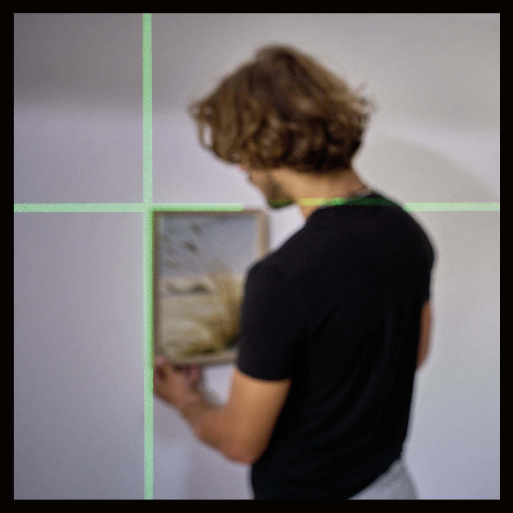 Eine Person mit braunen Haaren hält einen Bilderrahmen in einem Raum mit weißen Wänden und grünen Linien, die auf das Bild projiziert werden.