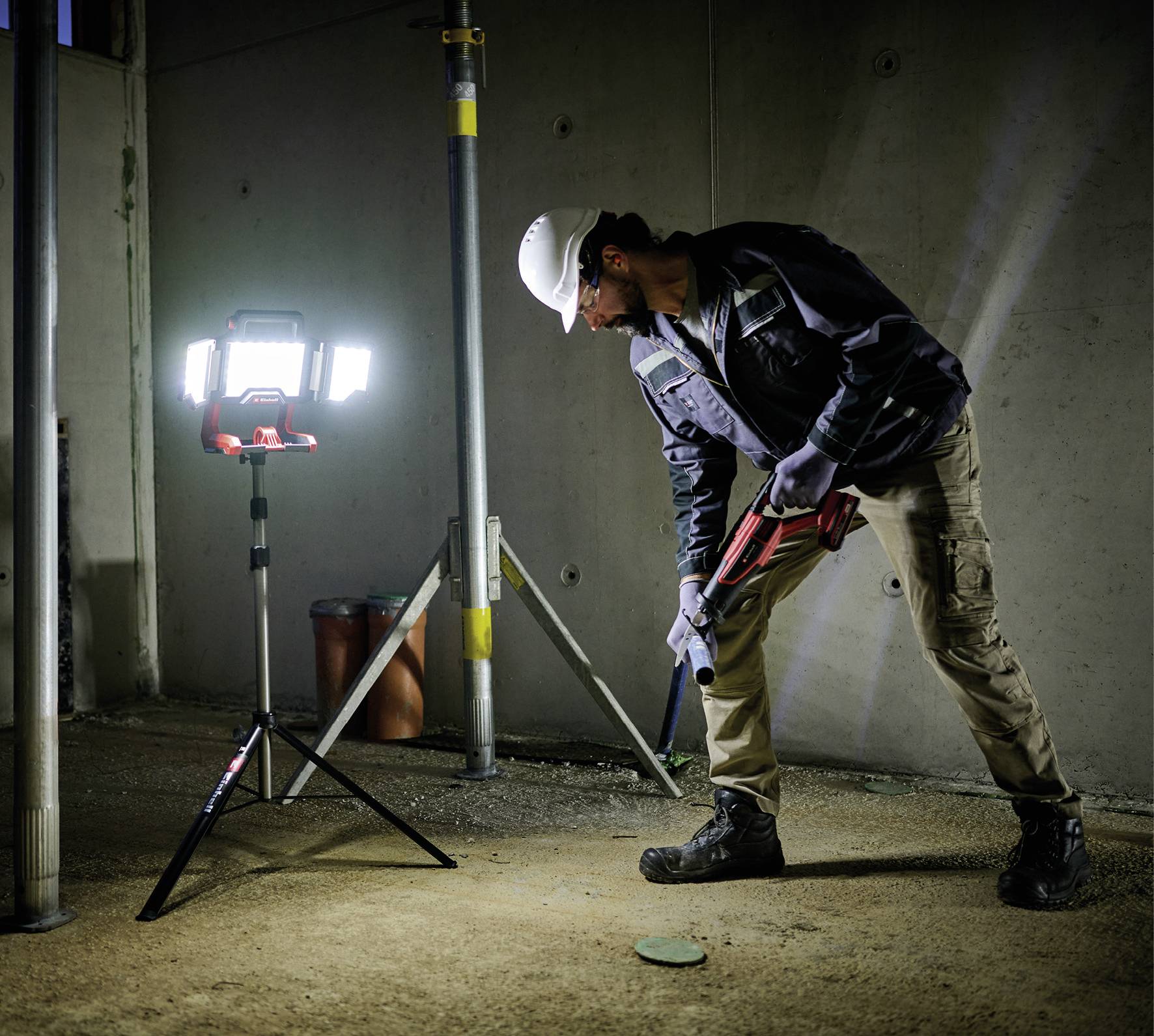 Eine Person arbeitet auf einer Baustelle mit einem Bohrhammer. Eine helle Baustellenlampe beleuchtet den Arbeitsbereich.