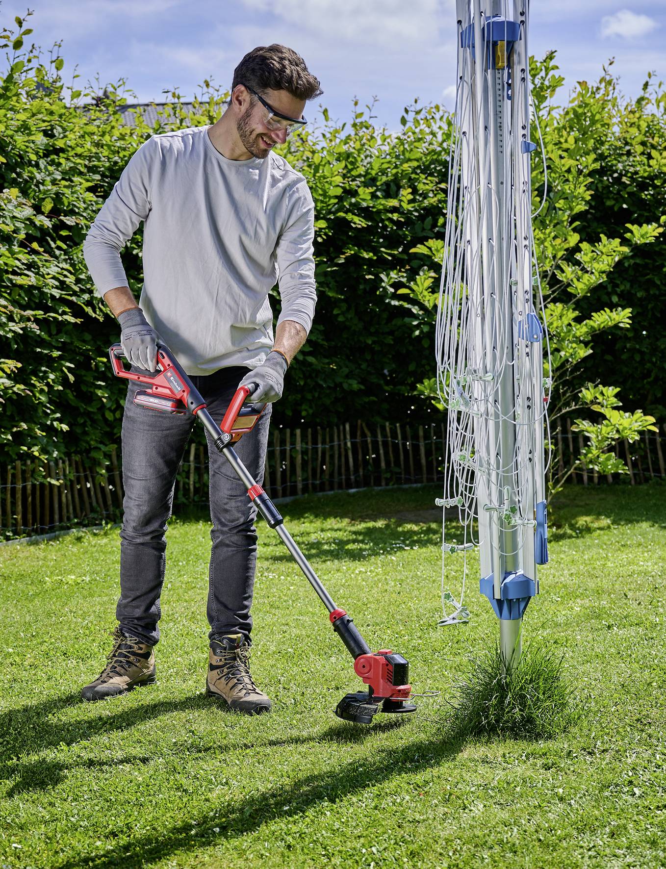 Eine Person trimmt den Rasen um einen Wäschespinner in einem Garten. Sie trägt Handschuhe und Schutzbrille, während sie arbeitet.