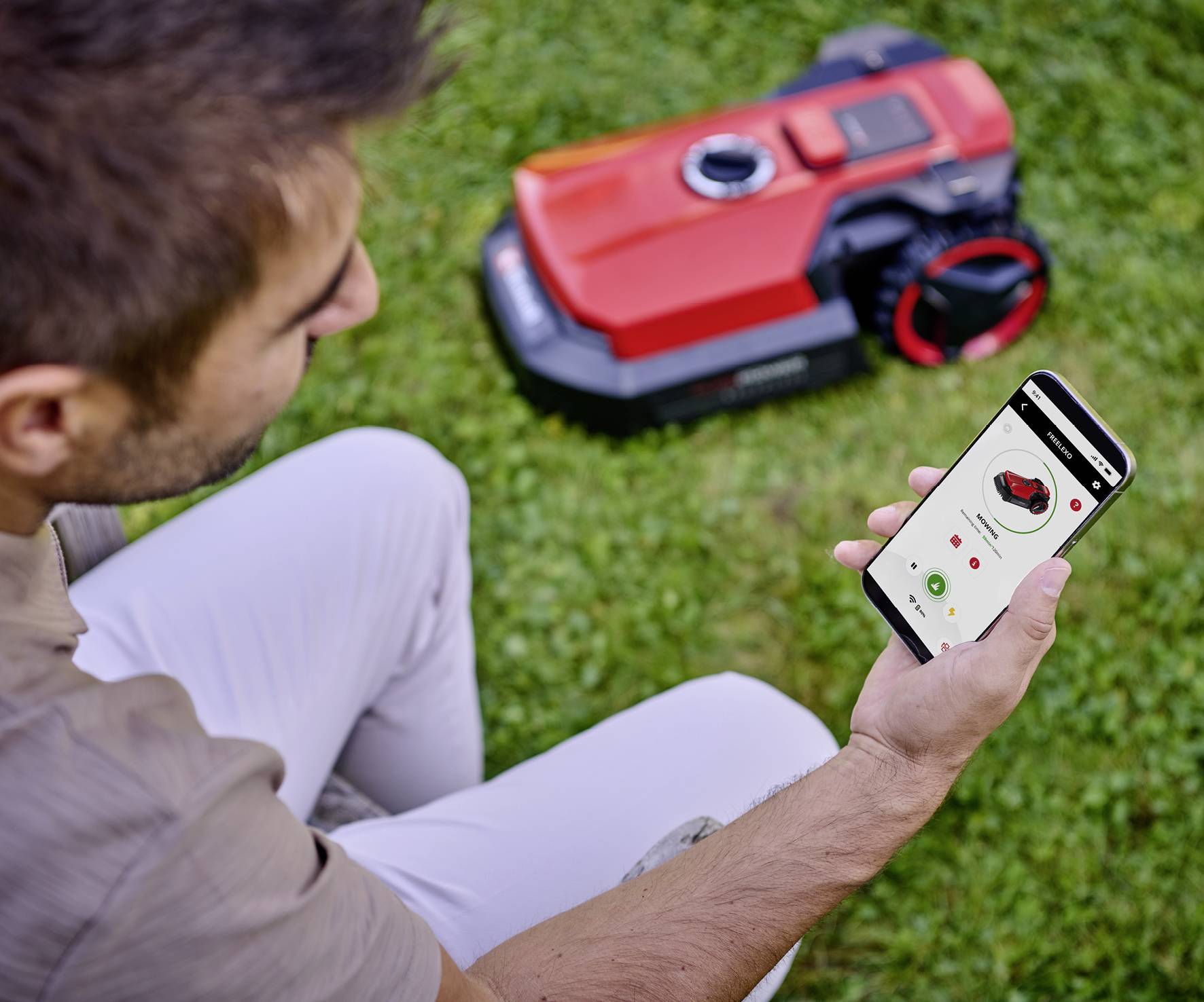 Ein Mann steuert einen roten automatischen Rasenmäher auf einer grünen Wiese mit einem Smartphone.