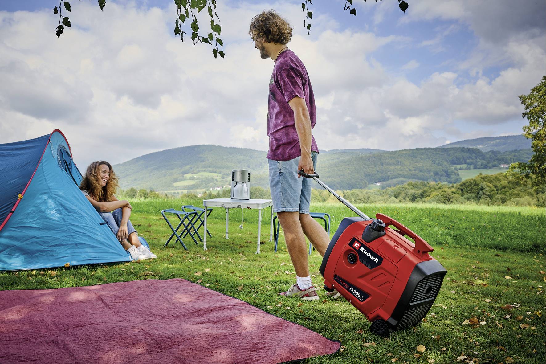 Mann zieht roten Generator auf einer Wiese beim Camping. Eine Frau sitzt neben einem Zelt. Im Hintergrund sind Hügel und ein Tisch.