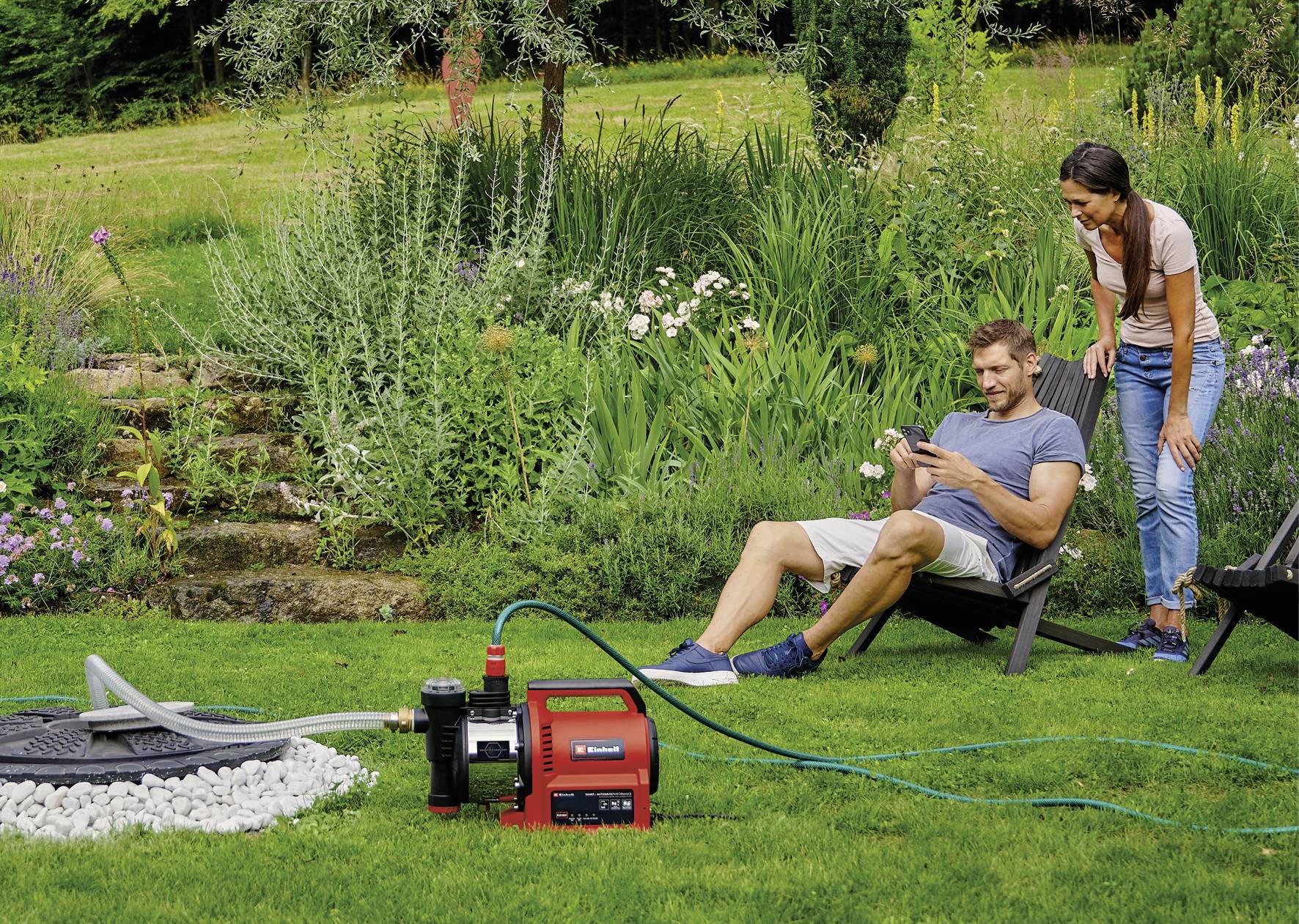 Ein Mann sitzt auf einem Liegestuhl im Garten und benutzt ein Smartphone. Eine Frau steht daneben und schaut auf das Display. Im Vordergrund ein Wasserpumpsystem.