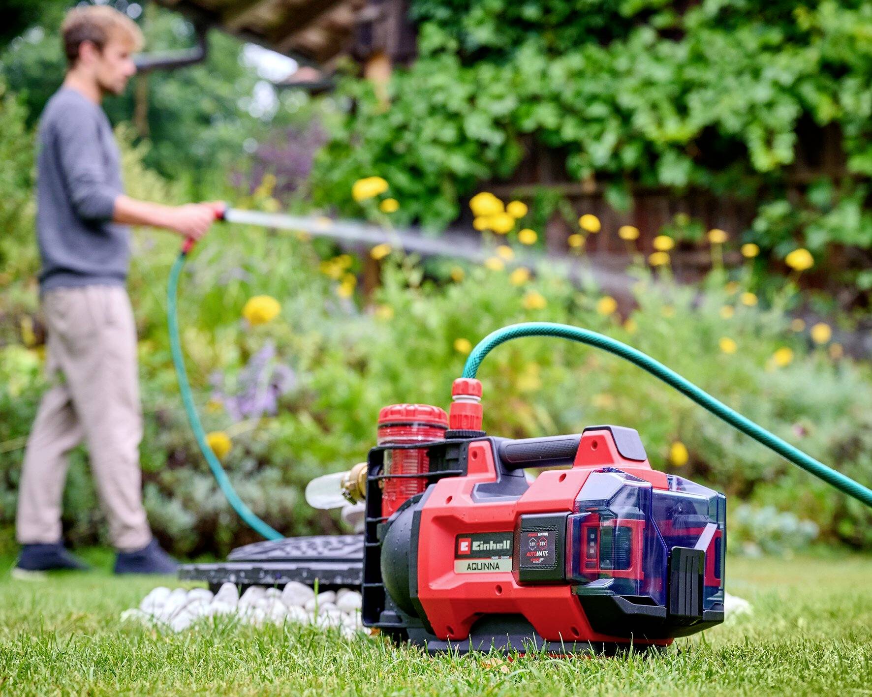 Ein Mann gießt mit einem Schlauch den Garten. Im Vordergrund befindet sich eine rote Wasserpumpe auf dem Rasen. Im Hintergrund sind Blumen sichtbar.