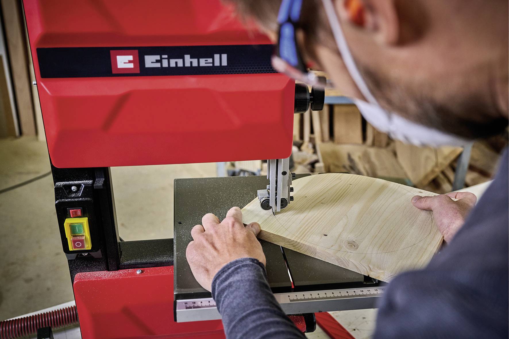 Eine Person schneidet ein Stück Holz mit einer roten Bandsäge der Marke Einhell in einer Werkstatt. Konzentration ist sichtbar.