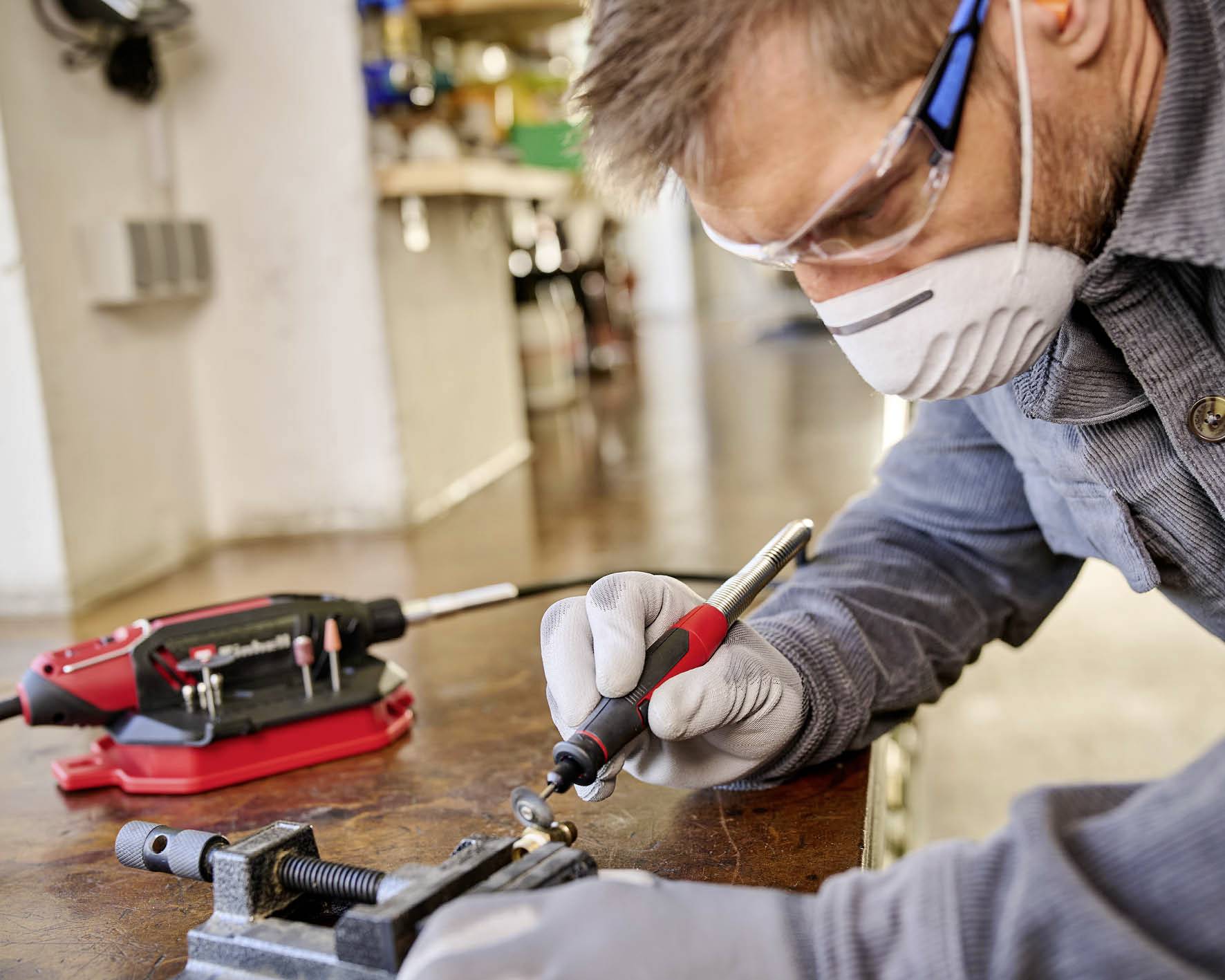 Ein Mann trägt Schutzbrille und Maske, während er mit einem Lötkolben an einem Metallstück arbeitet. Im Hintergrund sind Werkzeugkoffer und Werkstattutensilien zu sehen.