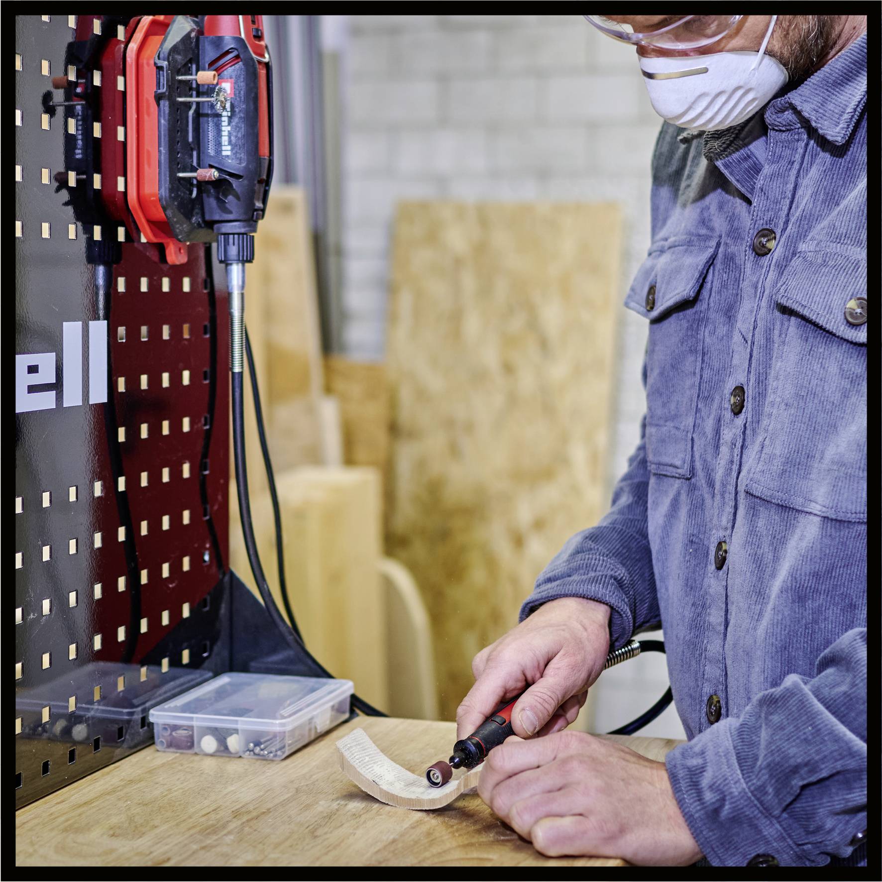 Eine Person trägt Schutzbrille und Atemschutzmaske, arbeitet an einem Holzstück mit einem elektrischen Werkzeug in einer Werkstatt.