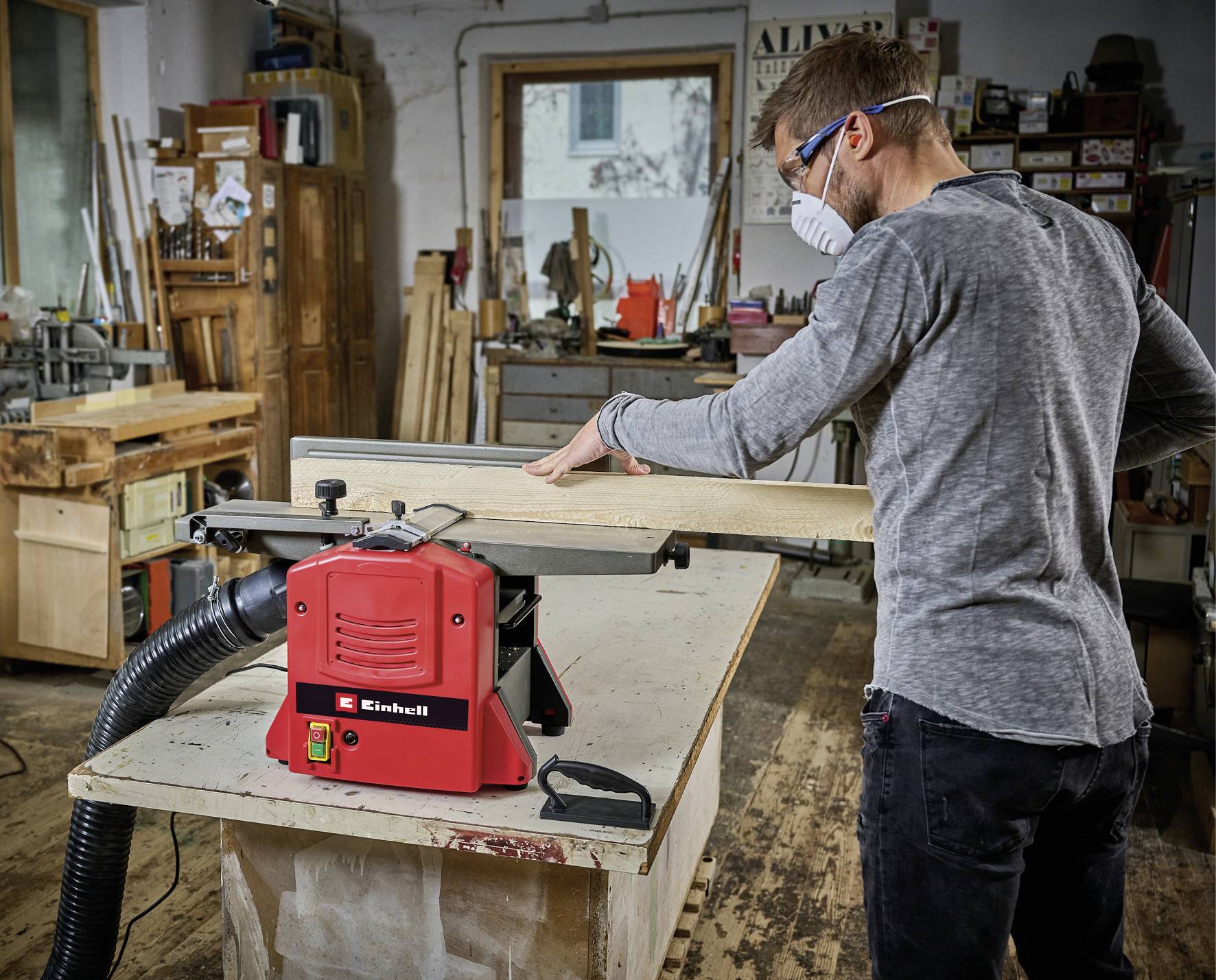 Ein Mann schleift ein Holzbrett mit einem Elektrowerkzeug in einer Werkstatt. Er trägt eine Schutzbrille und eine Maske.