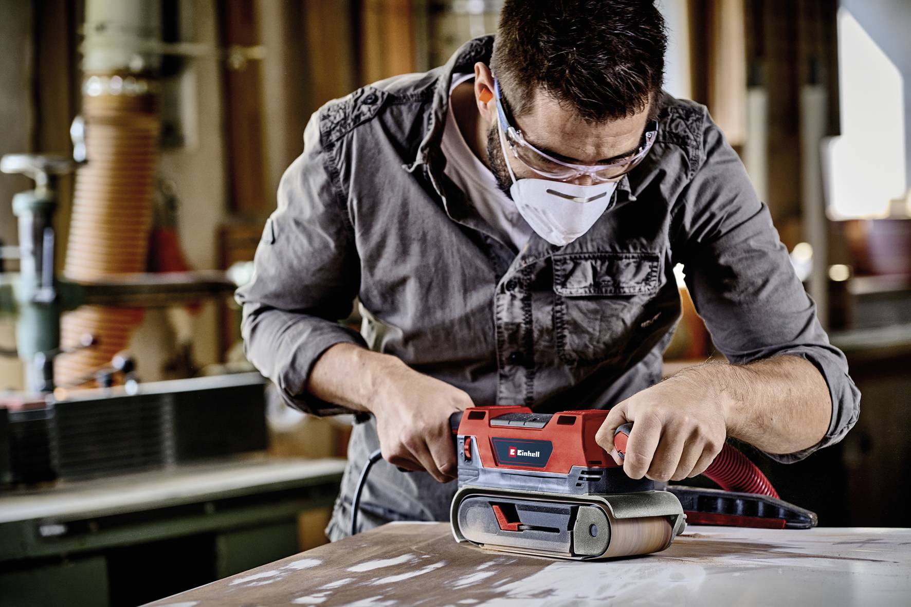Ein Mann schleift mit einem roten Bandschleifer Holz in einer Werkstatt. Er trägt Schutzbrille und Maske zum Schutz vor Staub.