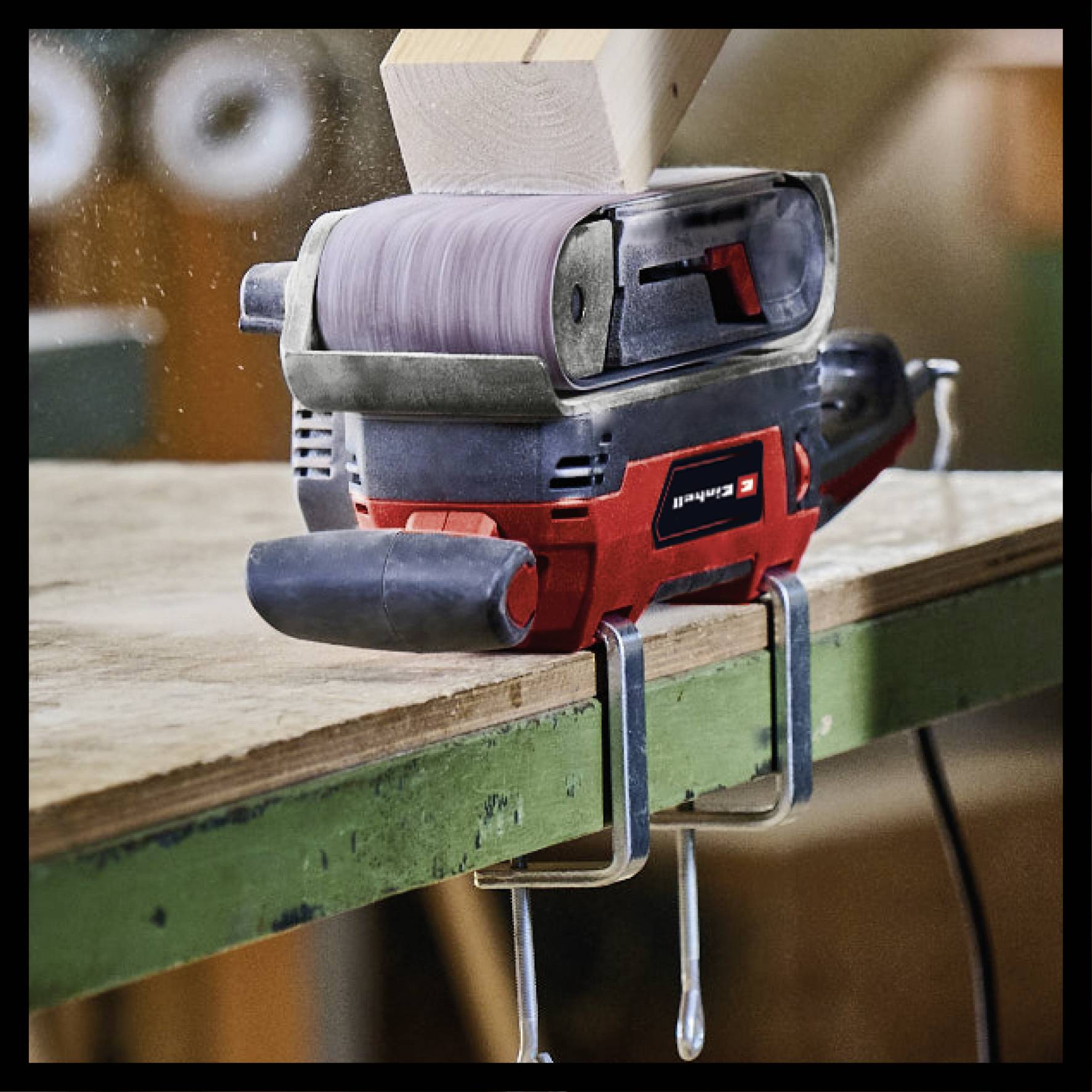 Schleifmaschine auf Arbeitstisch glättet Holzbalken. Maschine vibriert und erzeugt Staub. Hintergrund zeigt unscharfe Werkzeuge.