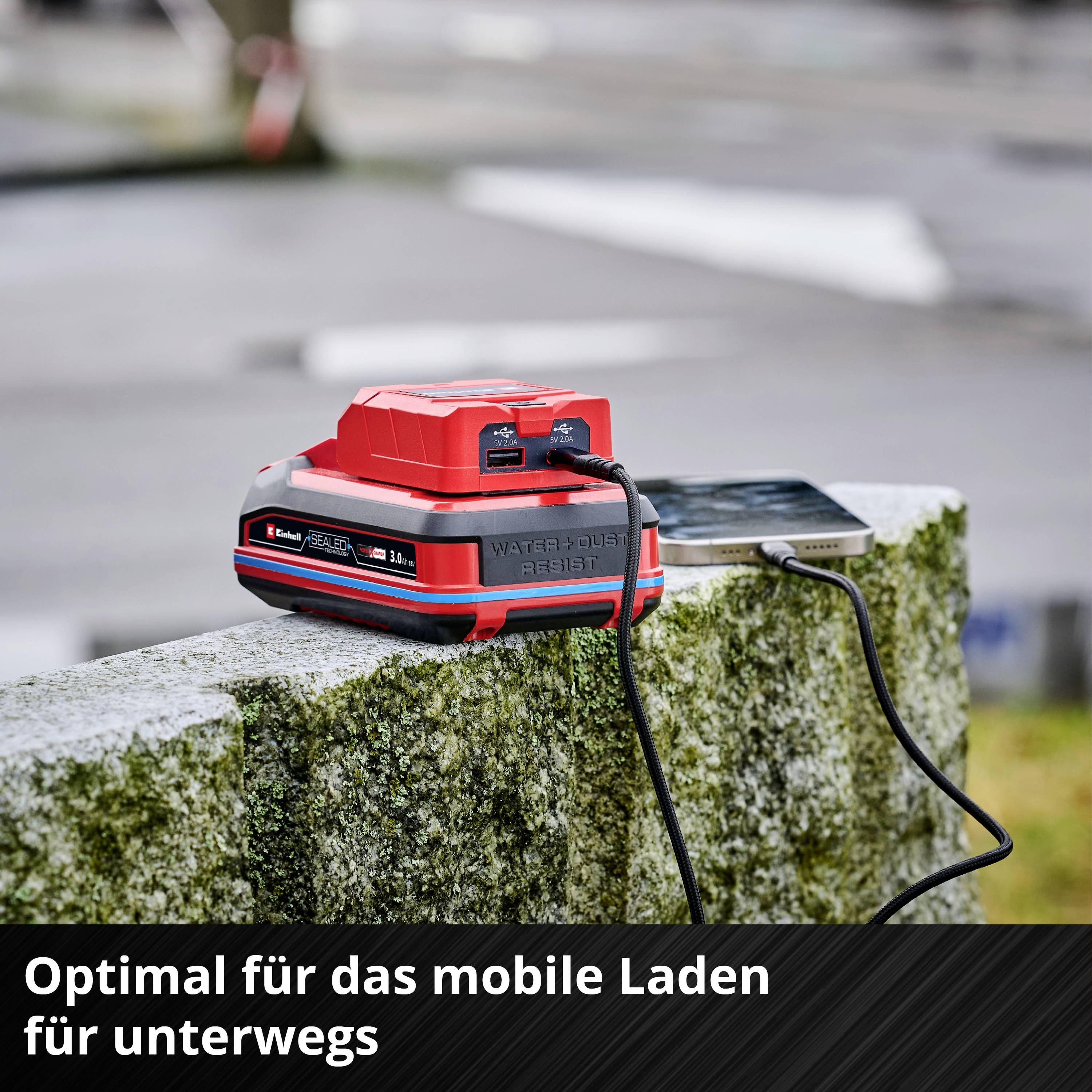 Ein tragbares Ladegerät lädt ein Smartphone im Freien auf. Der Text am unteren Rand lautet: 'Optimal für das mobile Laden für unterwegs'.