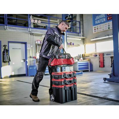 Ein Mann in Arbeitskleidung, zieht eine rote Werkzeugtasche auf einem stapelbaren Rollkoffer durch eine Werkstatt mit Hebebühnen und Werkzeugen.