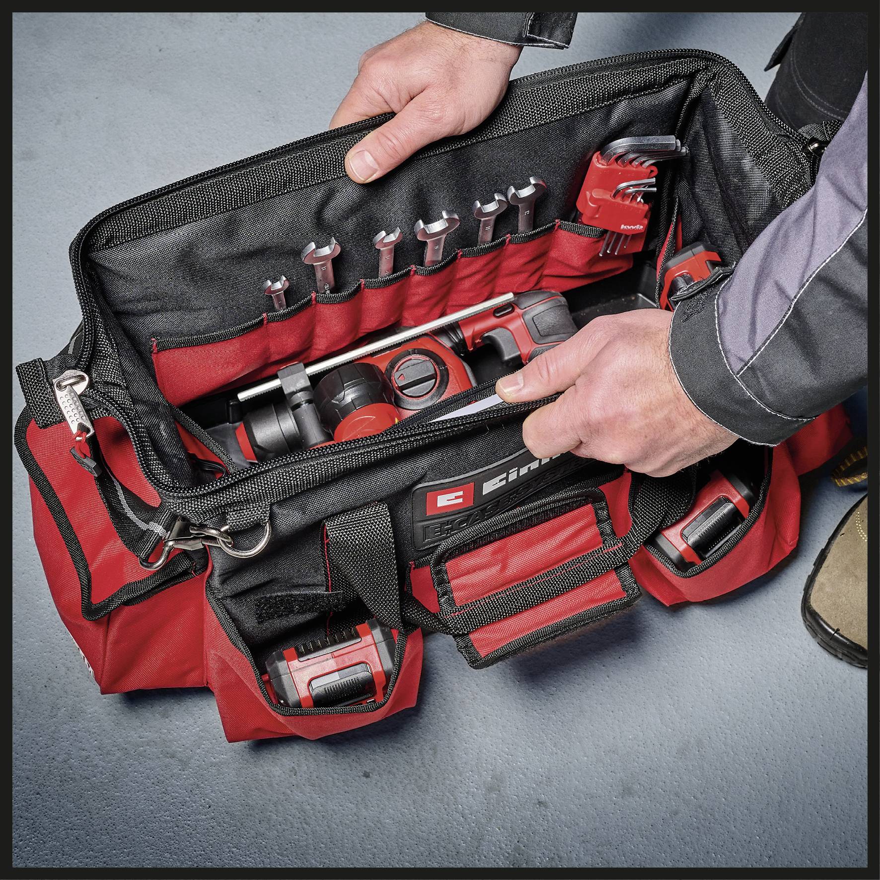 Eine rote Werkzeugtasche mit geöffnetem Reißverschluss zeigt verschiedene Werkzeuge, darunter Schraubendreher, Zangen und einen Hammer. Zwei Hände ordnen die Werkzeuge.
