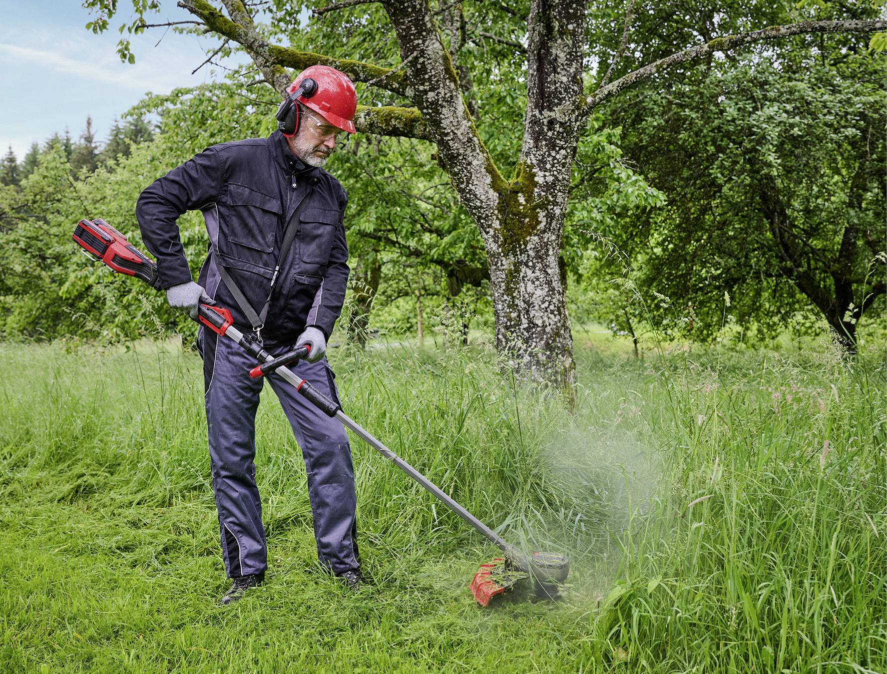 Eine Person mäht mit einem Freischneider Gras in einem grünen Garten, trägt Schutzkleidung und einen roten Helm, umgeben von Bäumen.