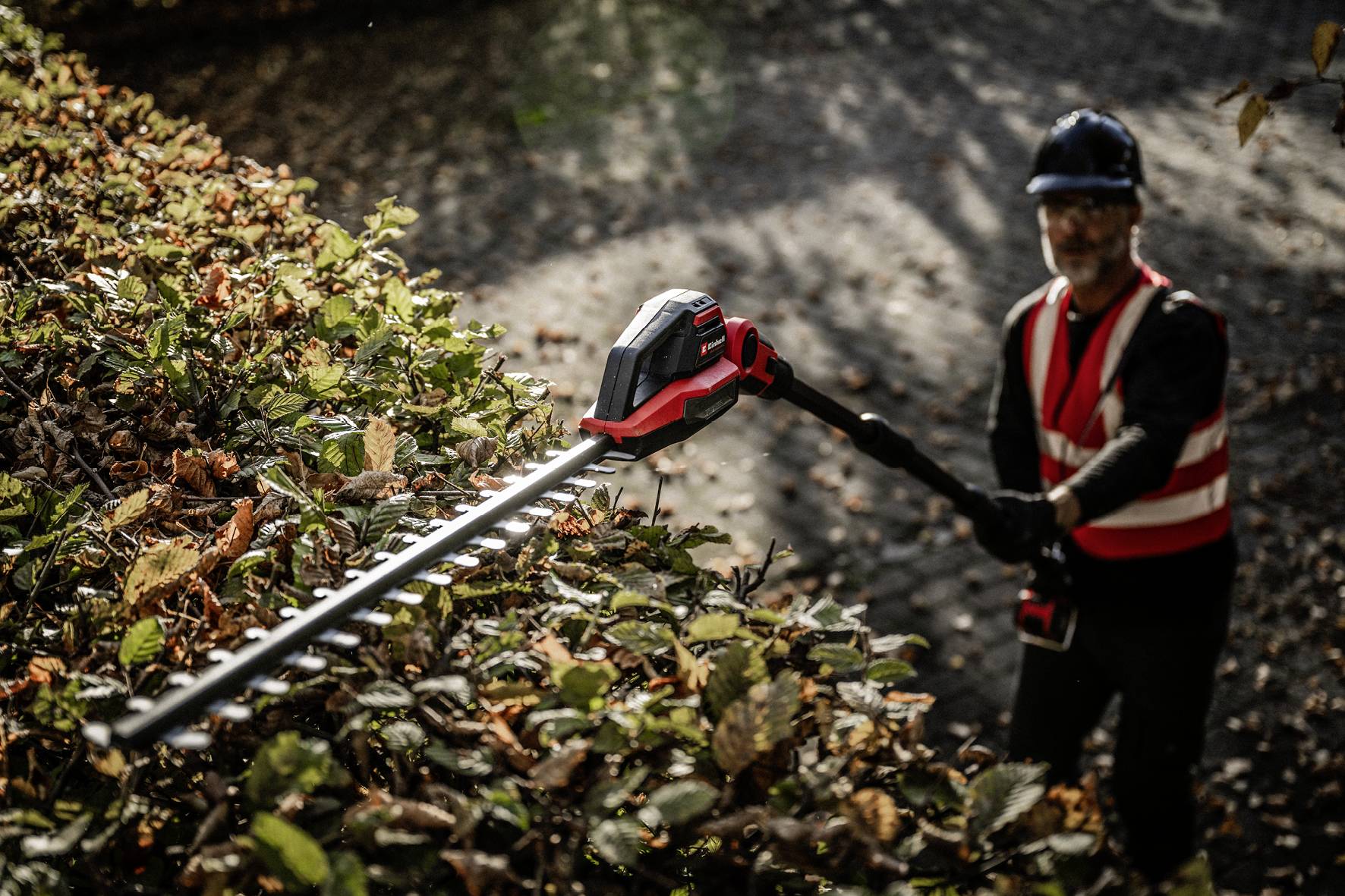 Person in Sicherheitskleidung schneidet Hecke mit elektrischer Heckenschere. Baumblätter im Vordergrund, sonniger Tag.