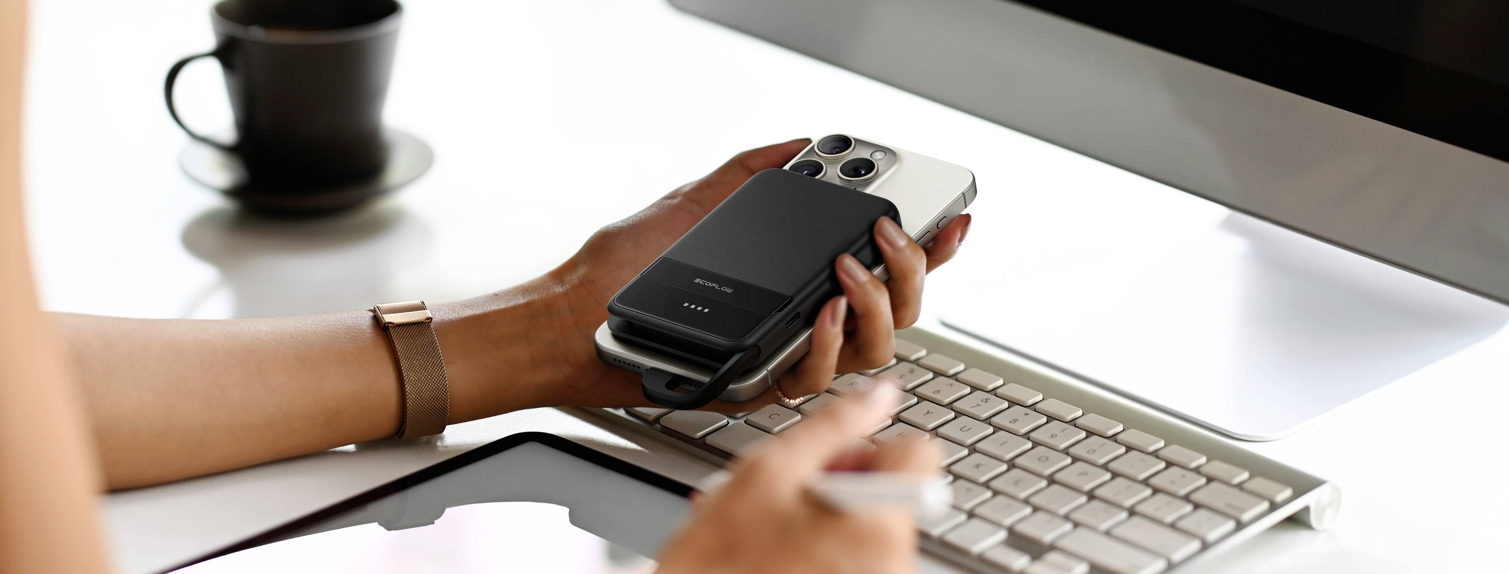 Eine Person hält ein Smartphone mit einem aufgesetzten Magnetladegerät über einer Computertastatur. Im Hintergrund steht eine Tasse.