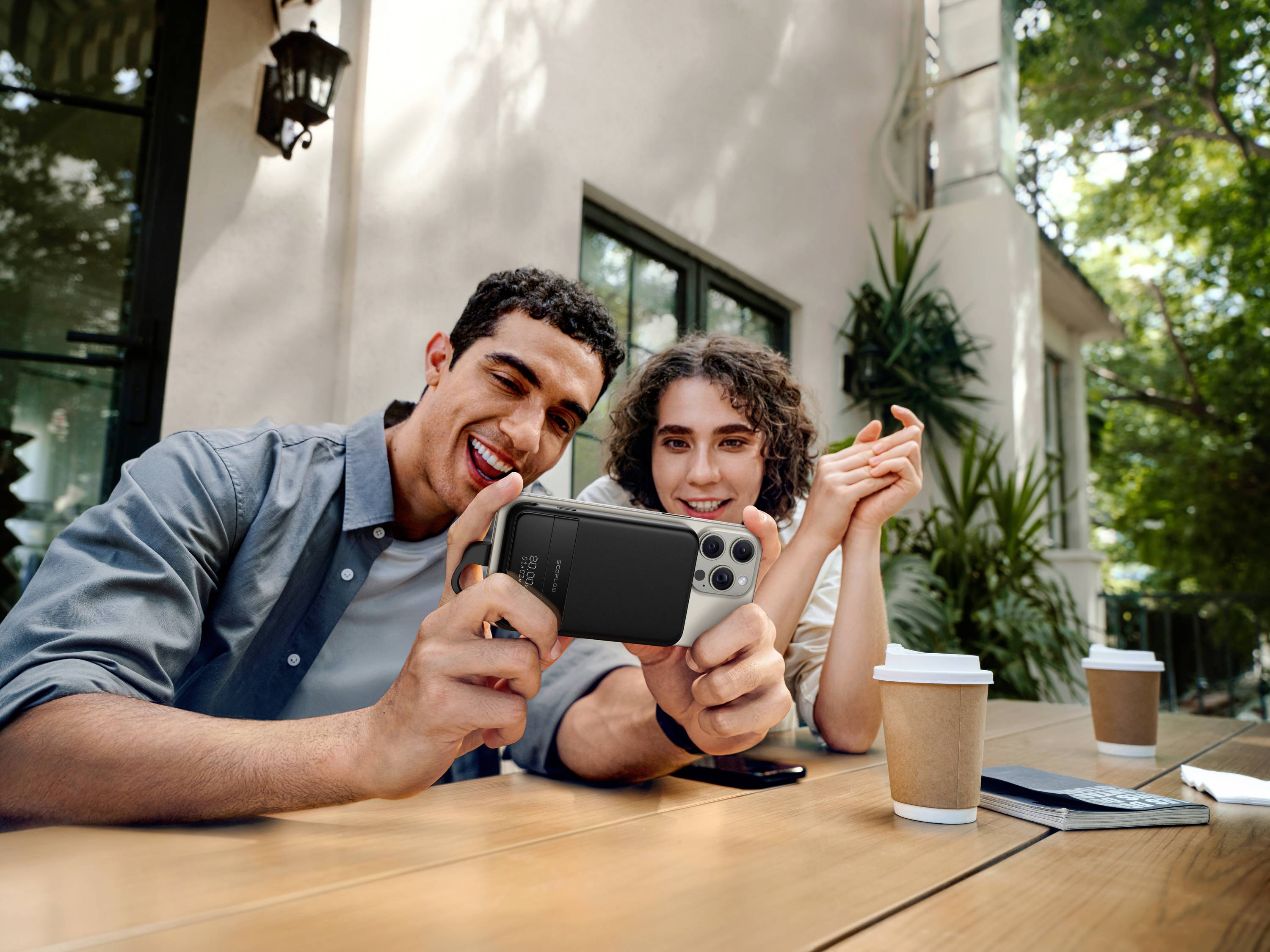 Ein Mann und eine Frau sitzen draußen an einem Tisch, lächelnd und betrachten ein Smartphone. Vor ihnen stehen zwei Pappbecher.
