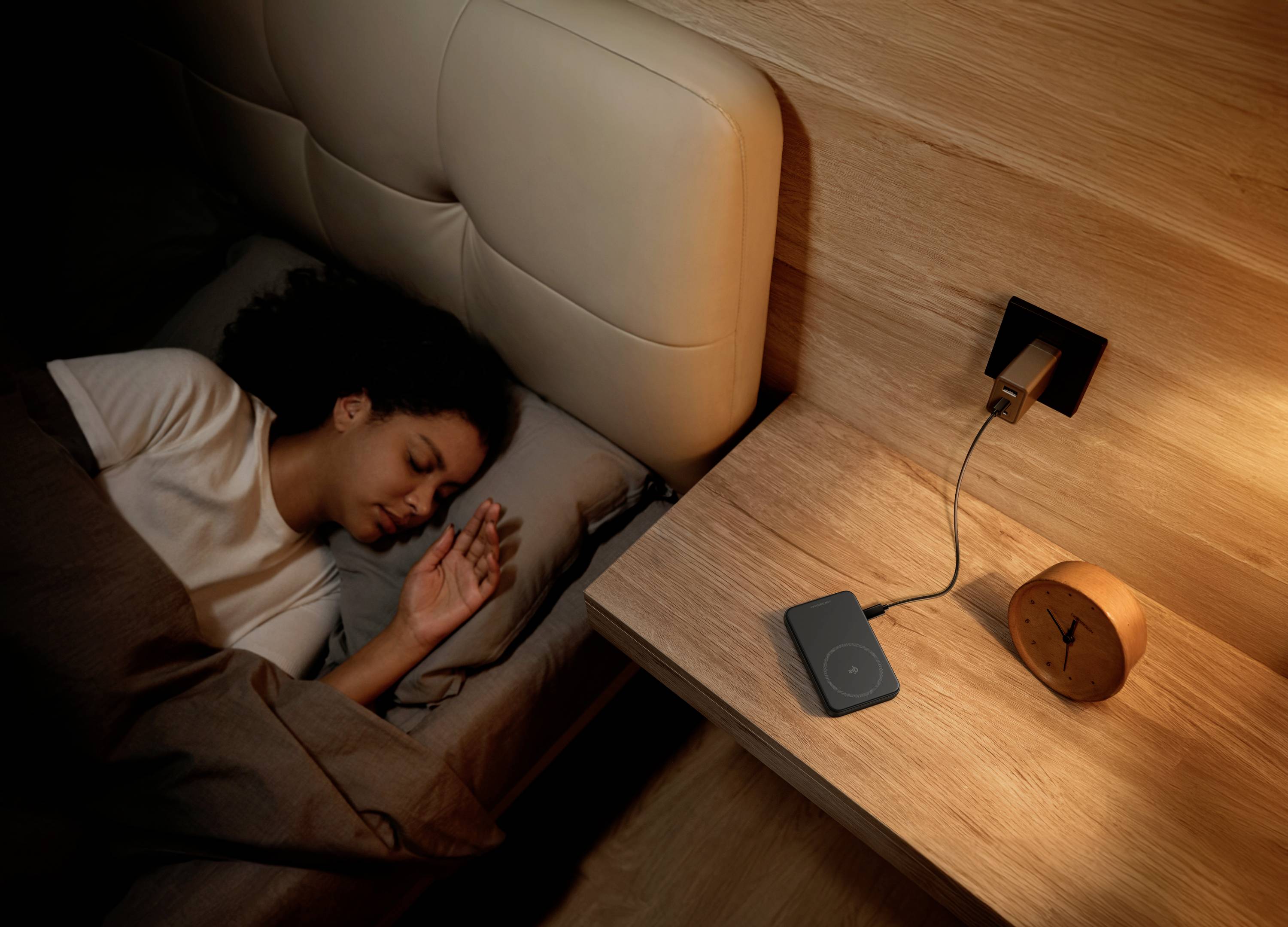 Eine schlafende Person im Bett neben einem Nachttisch mit einer Lampe, einem Wecker und einem Ladegerät. Gemütliche und ruhige Atmosphäre.