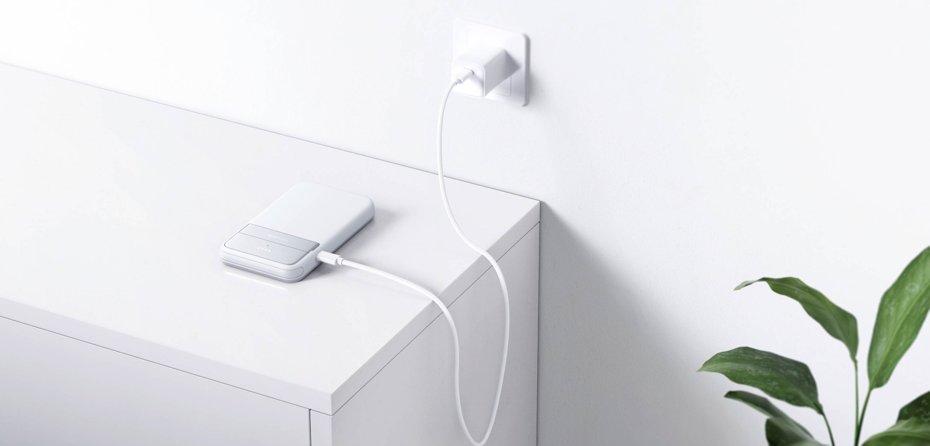 Ein Smartphone liegt auf einem weißen Tisch und lädt über ein Kabel, das mit einer Steckdose verbunden ist. Eine Pflanze steht daneben.