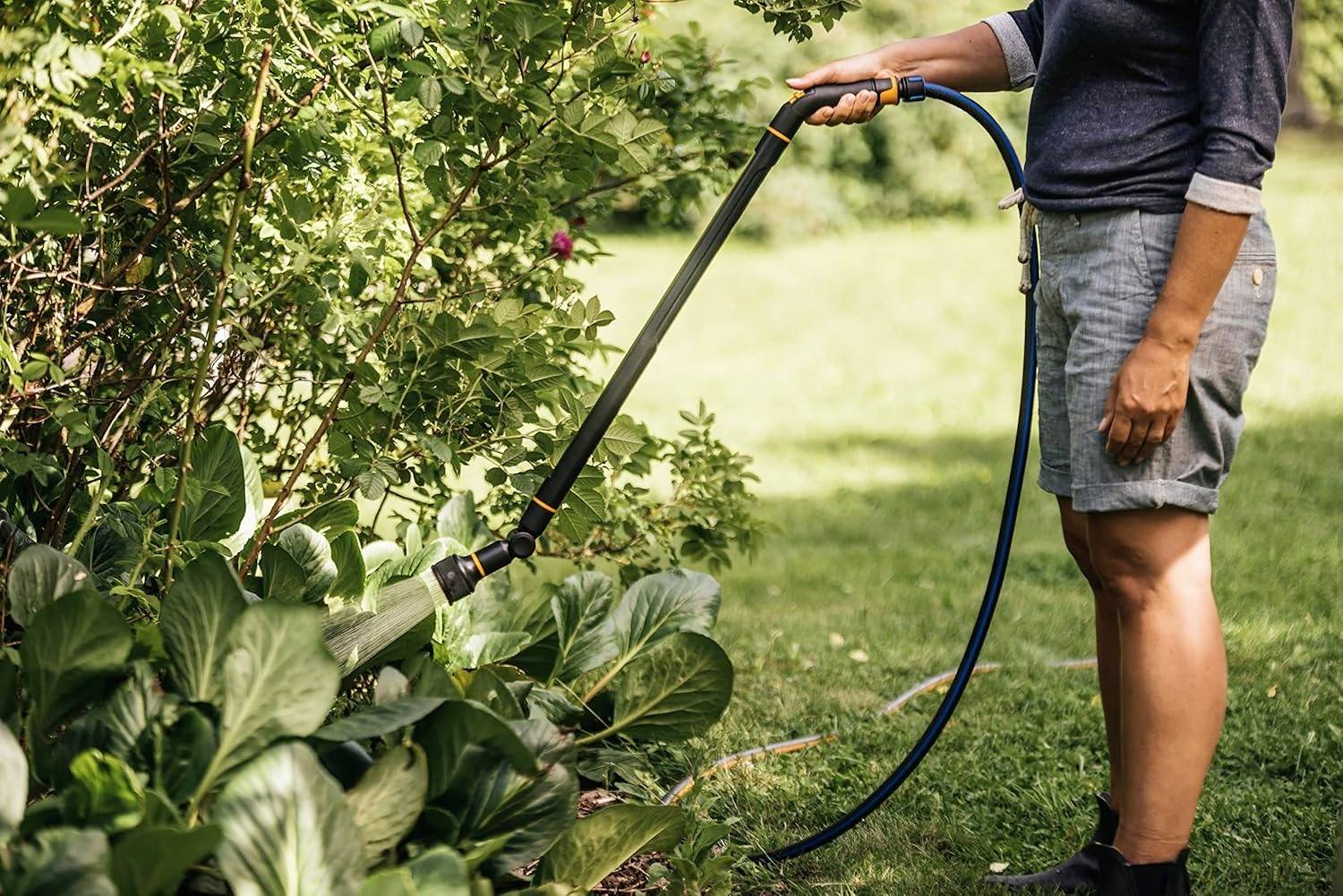 Eine Person bewässert einen Garten mit einem Schlauch, umgeben von grünen Pflanzen. Die Szene deutet auf Gartenarbeit hin.