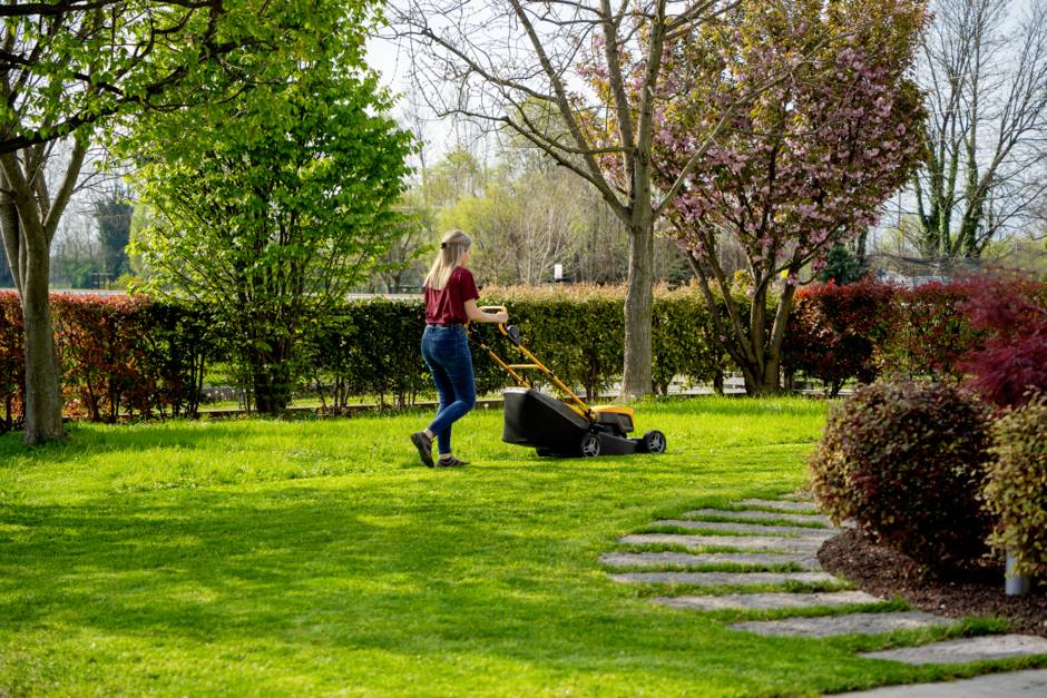 Person mäht den Rasen mit einem Rasenmäher in einem Garten mit blühenden Bäumen und gepflegtem Grün.