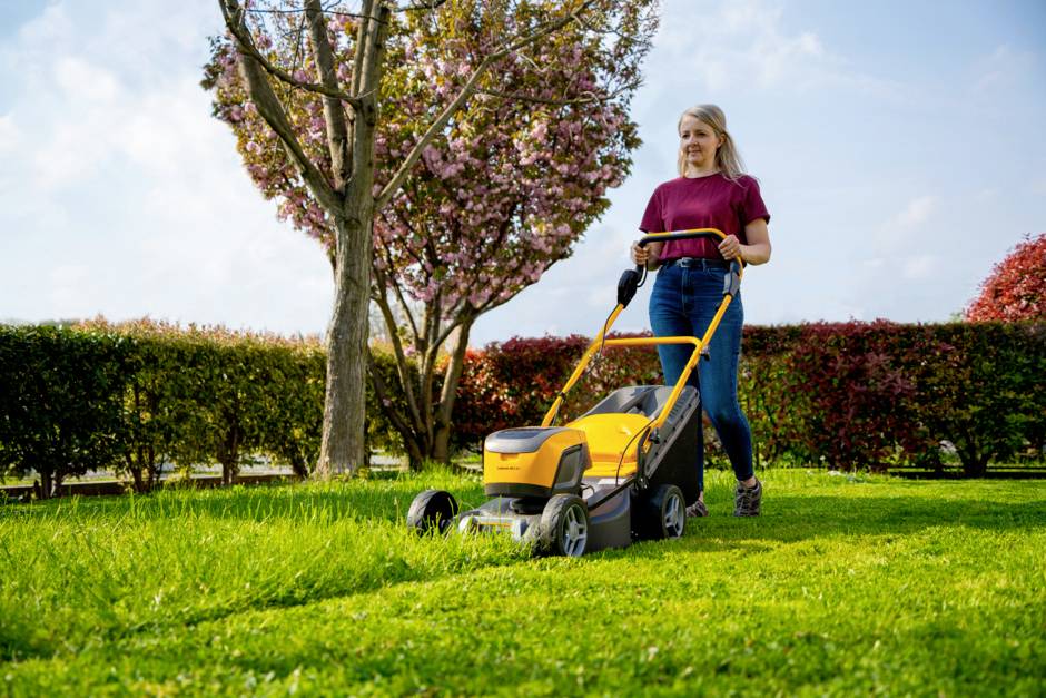 Eine Frau mäht den Rasen mit einem gelben Rasenmäher in einem Garten. Im Hintergrund blüht ein Baum, und eine Hecke ist sichtbar.
