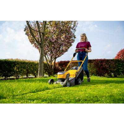 Eine Frau mäht den Rasen mit einem gelben Rasenmäher in einem Garten. Im Hintergrund blüht ein Baum, und eine Hecke ist sichtbar.