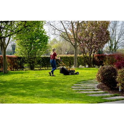 Eine Person mäht den Rasen in einem Garten mit einem Handrasenmäher. Im Hintergrund sind blühende Bäume und Sträucher zu sehen.