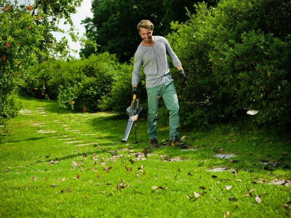 Mann benutzt Laubbläser, um Herbstblätter auf einem Gehweg in einem grünen Garten zu entfernen.