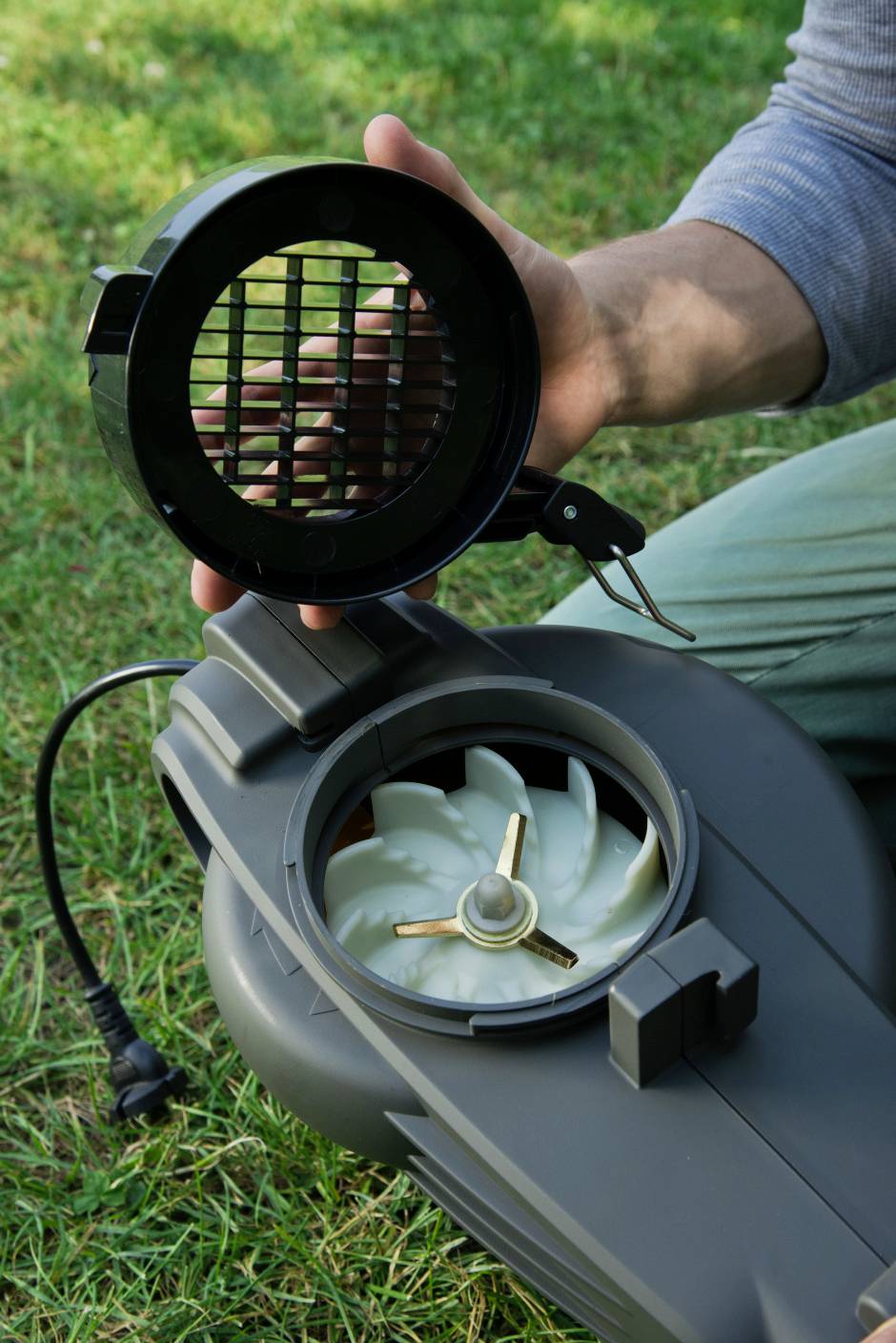 Eine Person hält den Deckel eines elektrischen Gartenhäckslers, der das Innenleben mit einem spiralförmigen Messer zeigt. Gerät steht auf Gras.