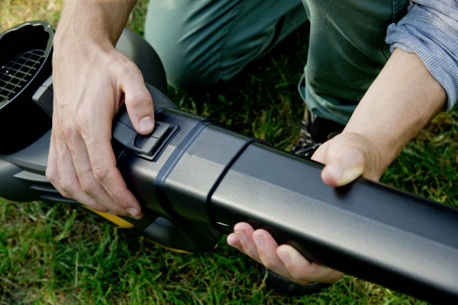 Eine Person kniet auf dem Rasen und montiert einen Laubbläser. Hände justieren Drehknopf am Gerät, das für Gartenarbeit genutzt wird.