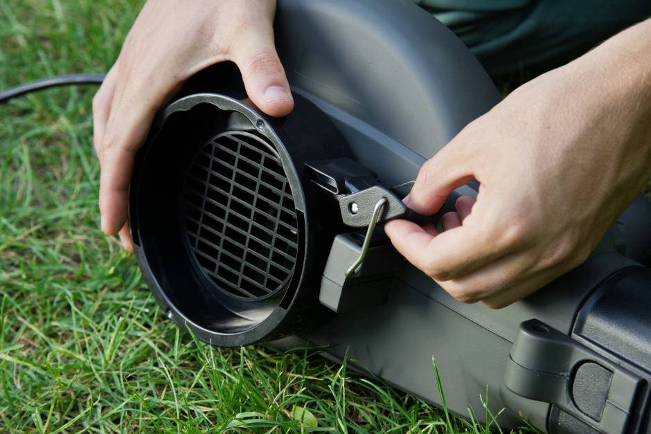 'Person hält und bedient ein elektrisches Gebläse im Gras. Hände positionieren einen Hebel am Gerät.'