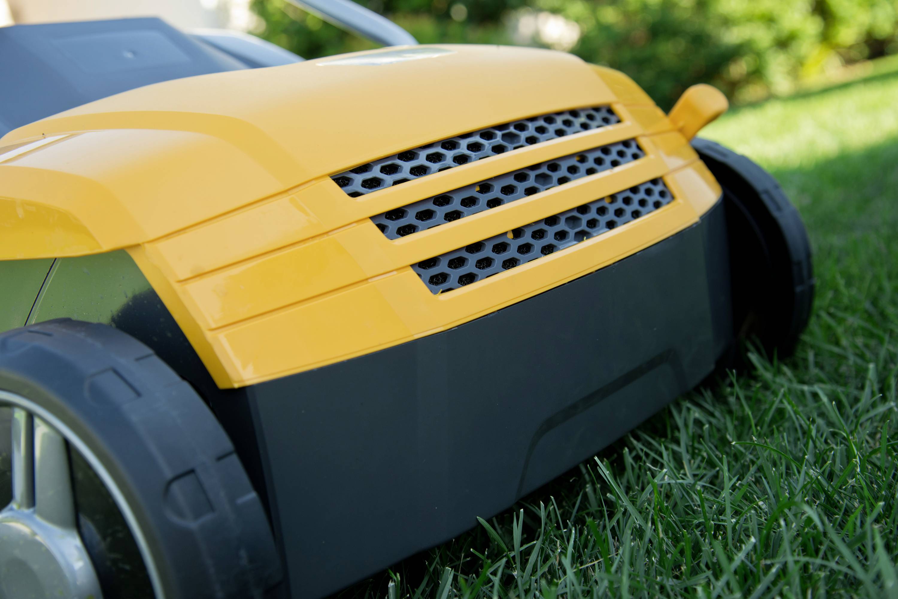 Ein orange-gelber Spielzeug-Rasenmäher steht auf grünem Gras. Vorderansicht zeigt Details der Lüftungsschlitze und Räder.