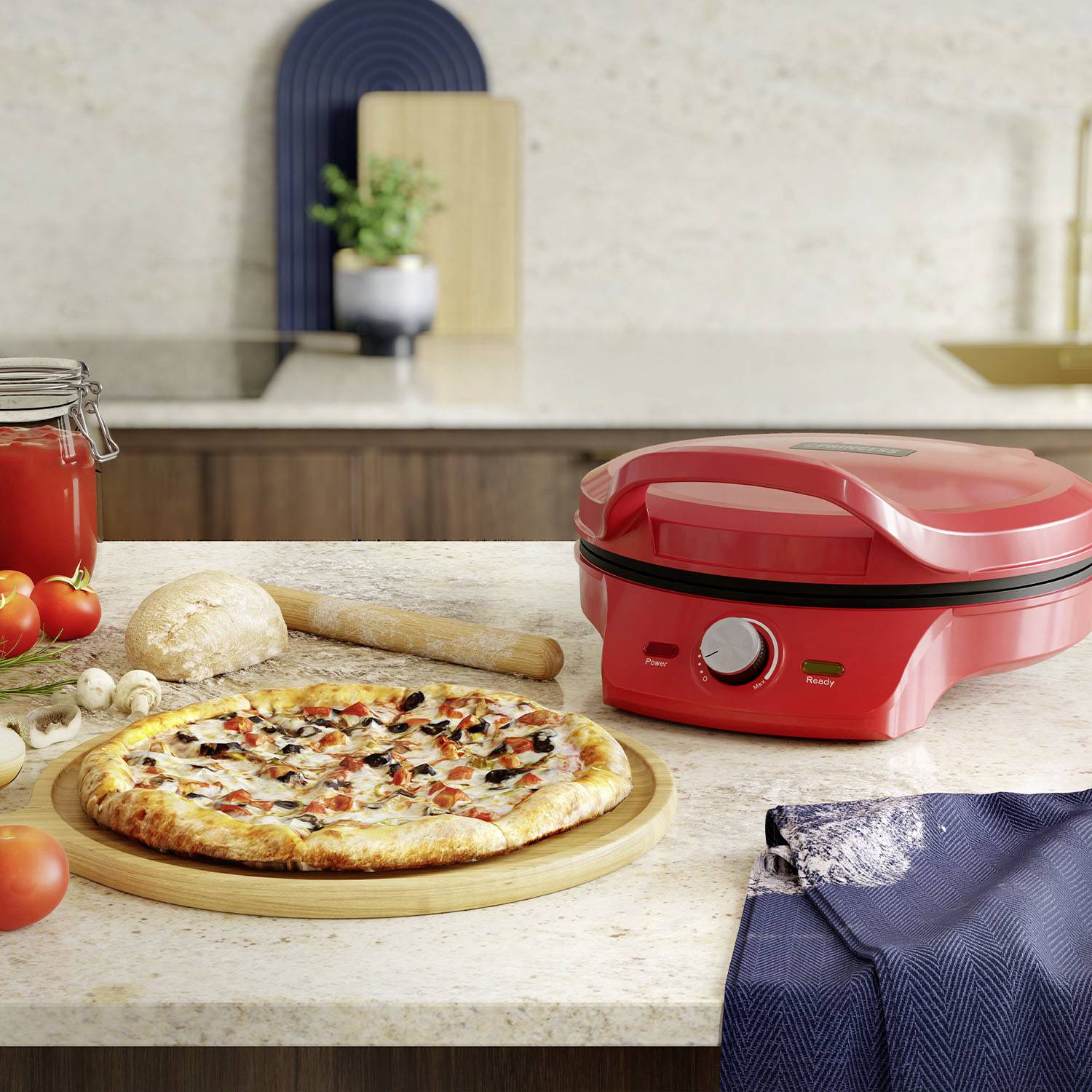 Ein roter Pizzamaker steht auf einer Küchenarbeitsplatte mit einer frisch gebackenen Pizza. Daneben liegen Zutaten wie Tomaten und Pilze.
