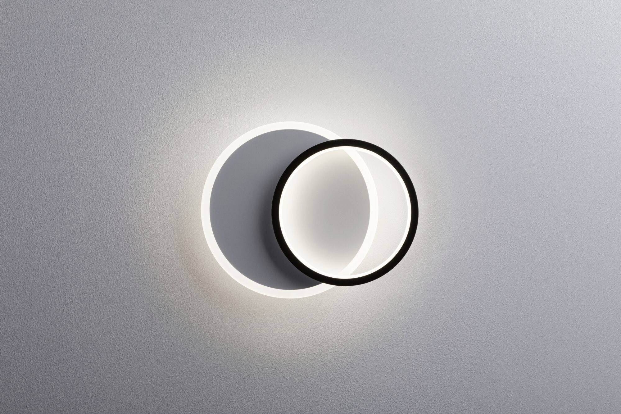 Kreisförmige Leuchte an der Wand, bestehend aus einem beleuchteten Ring und einem schwarzen Ring, die sich überlagern und einen Schatten werfen.