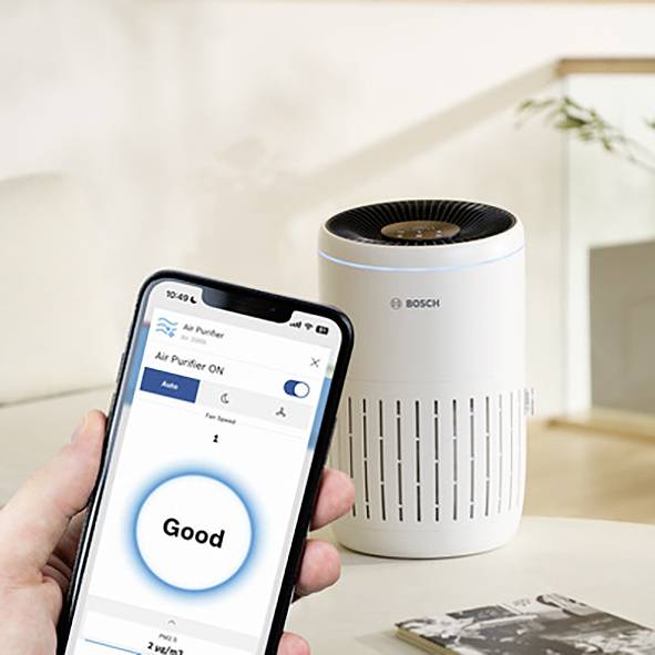 Ein Luftreiniger von Bosch steht auf einem Tisch. Daneben hält eine Hand ein Smartphone, das die Luftqualität als 'Good' anzeigt.