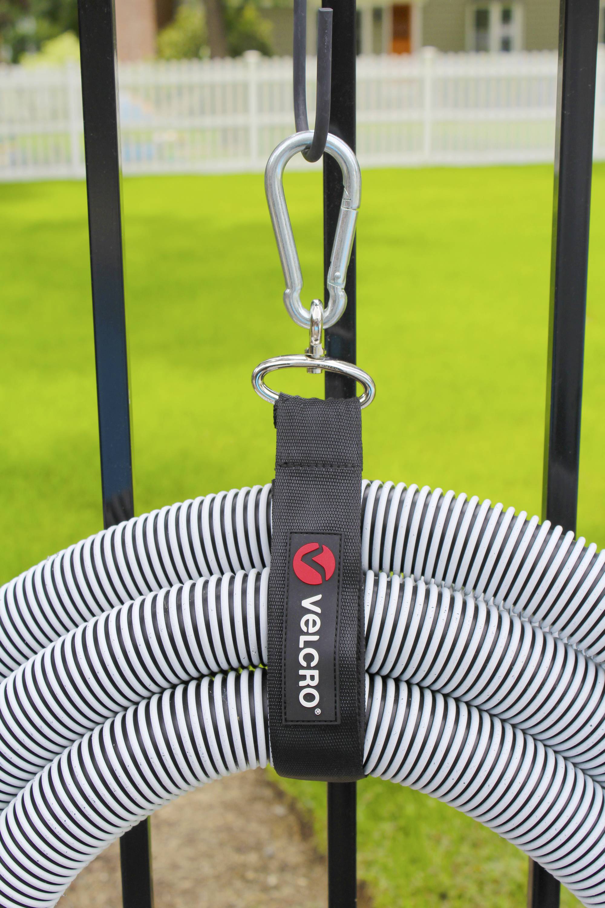 Ein weißer, spiralförmiger Schlauch wird an einem schwarzen Zaun mit einem Klettband und einem Karabinerhaken befestigt. Im Hintergrund ist Gras sichtbar.