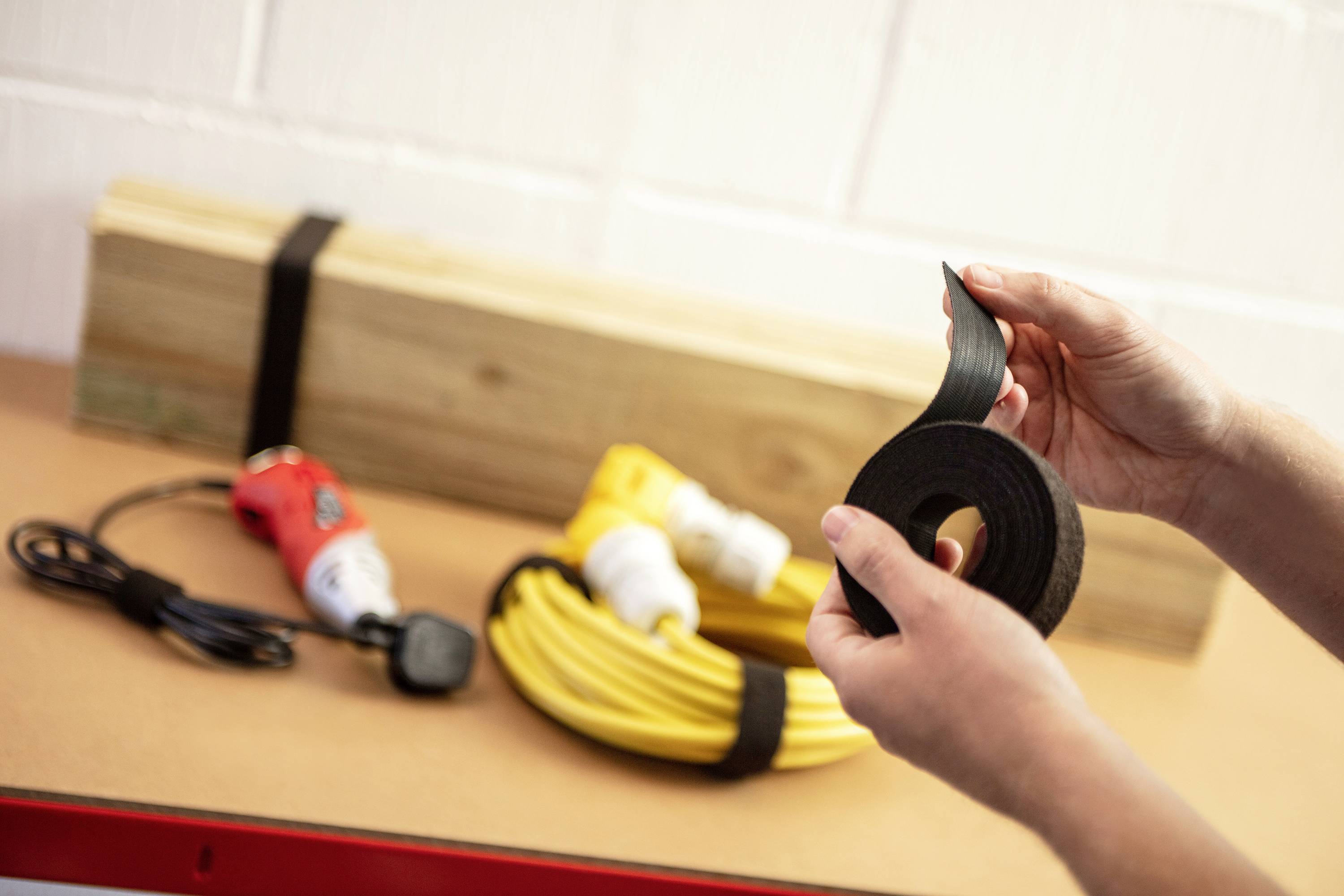 Person hält eine Rolle Klettband vor einem Hintergrund mit Holzlatten, Kabel und Bohrmaschine, die mit demselben Band zusammengebunden sind.